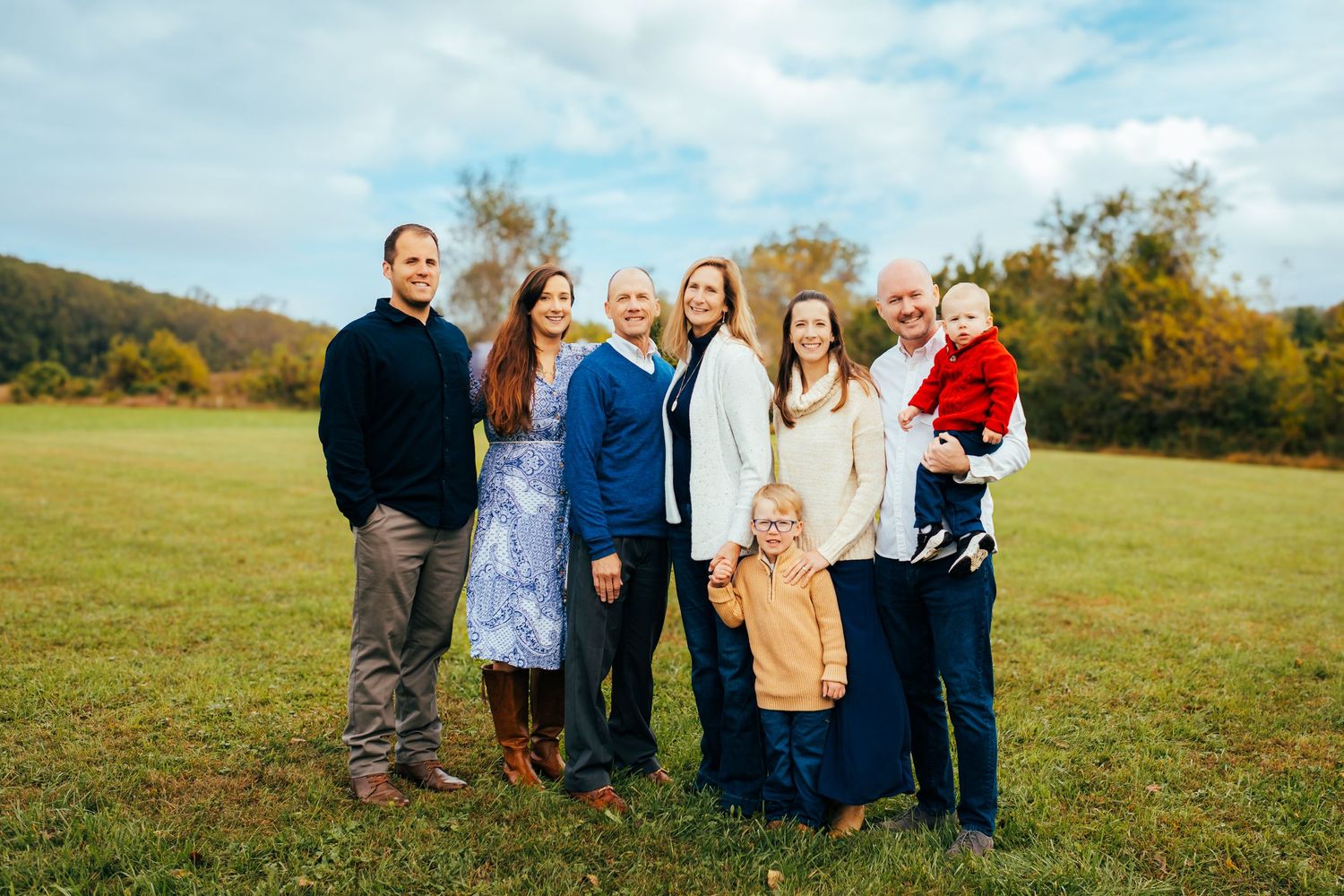 Large family group portrait outdoors in autumn field with colorful fall foliage and blue sky backdrop.
