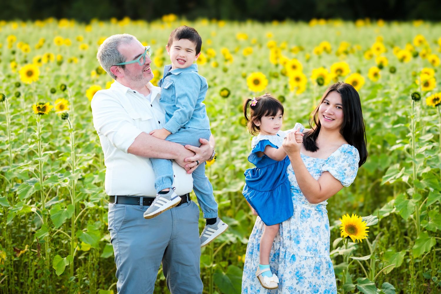Family posing in sunflower field, two adults holding young children, smiling outdoors on a sunny day.