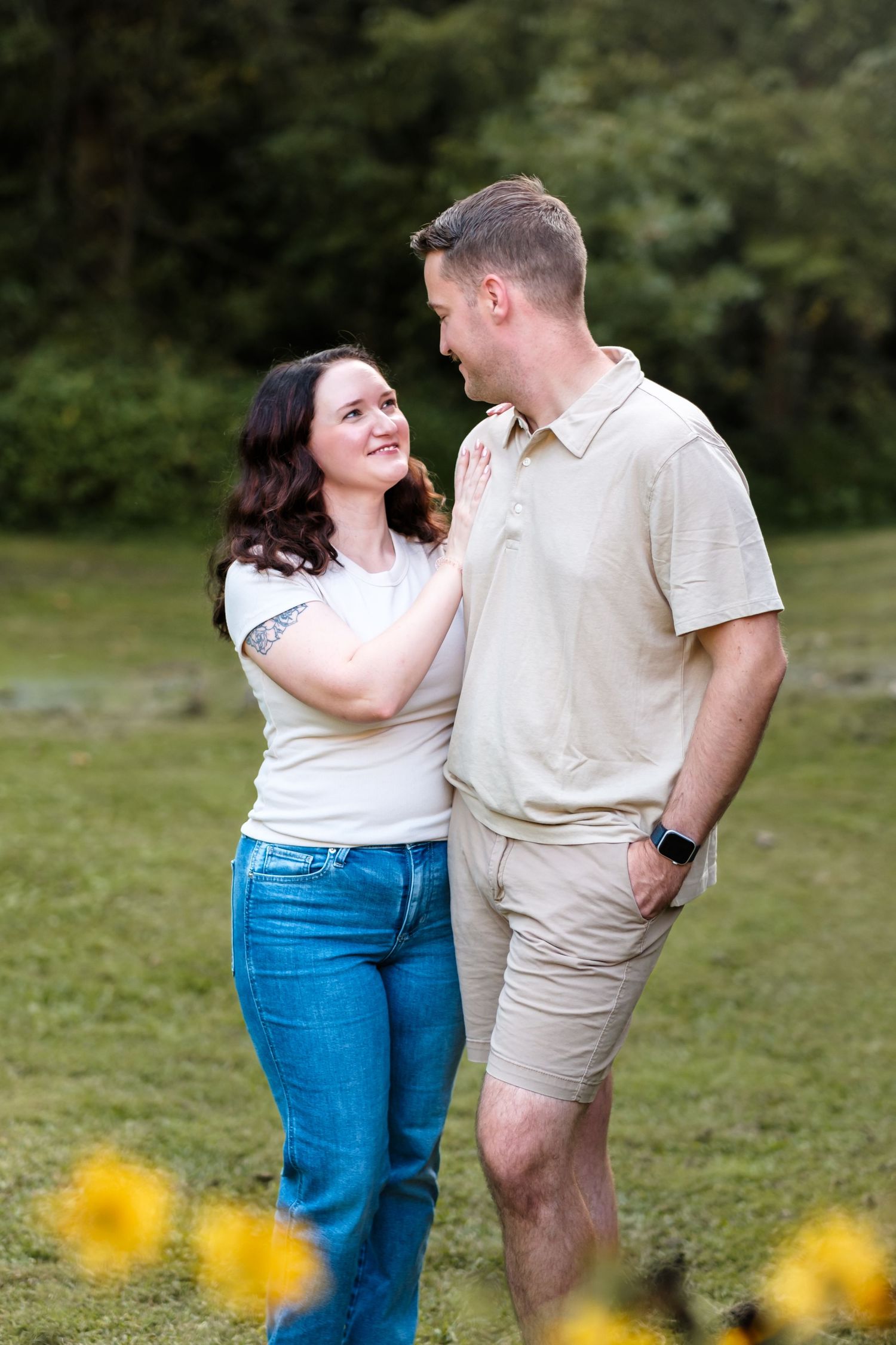 A couple smiles at each other outdoors, woman in white tee and jeans, man in beige polo and shorts, green park background.