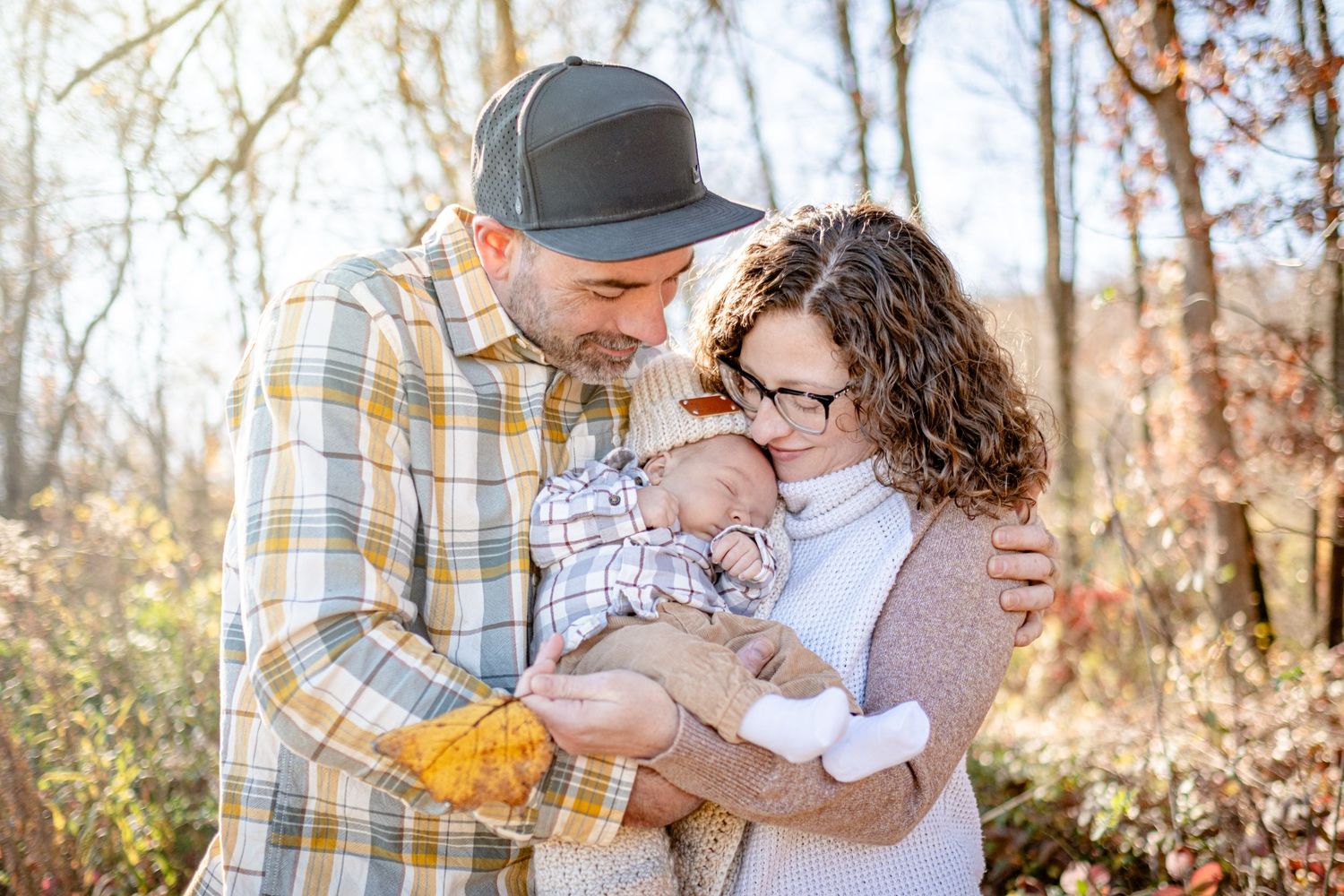 A couple tenderly holds their newborn baby outdoors among autumn trees in soft natural light.