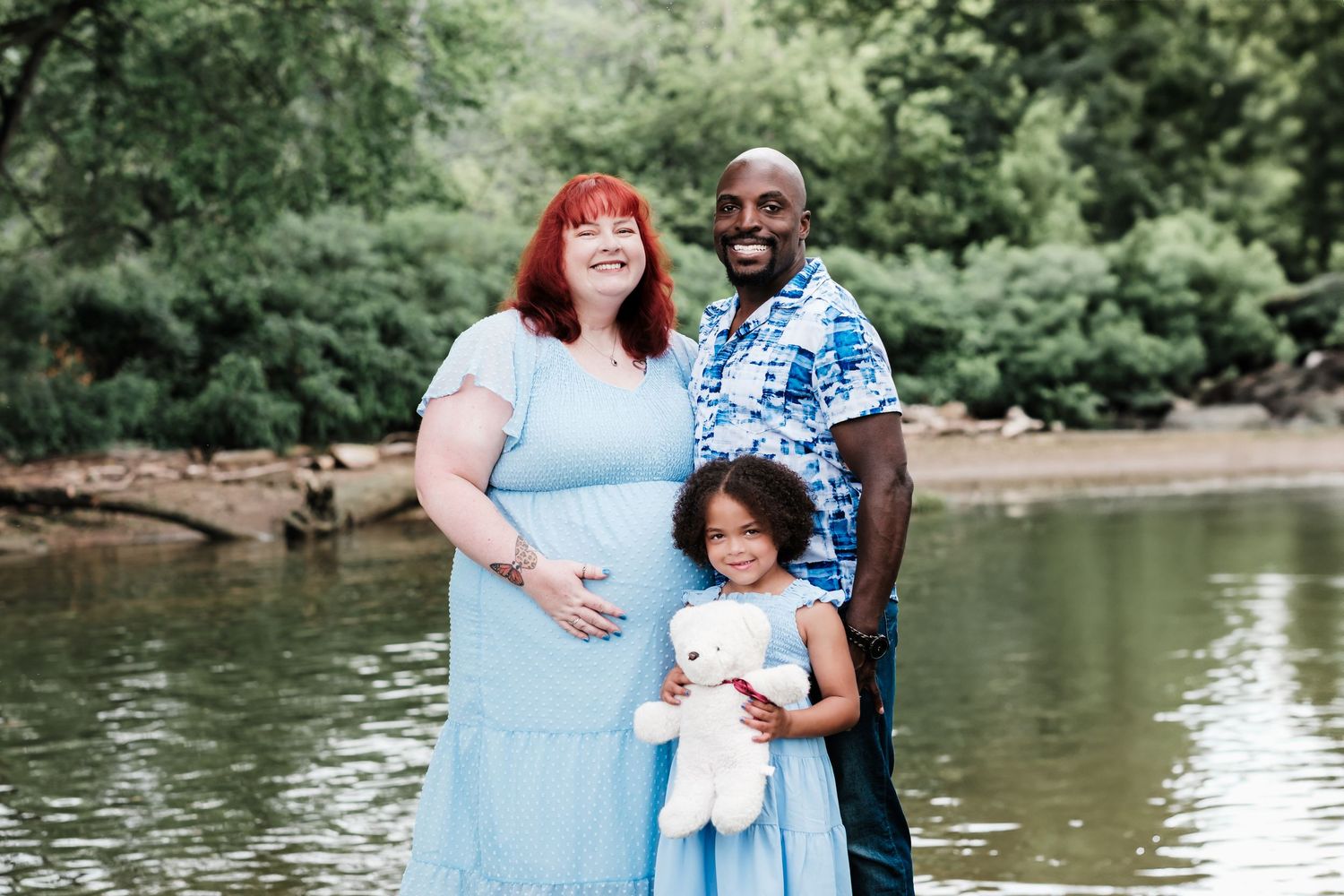 Pregnant woman in blue dress with husband and daughter holding teddy bear by a peaceful river surrounded by lush greenery.
