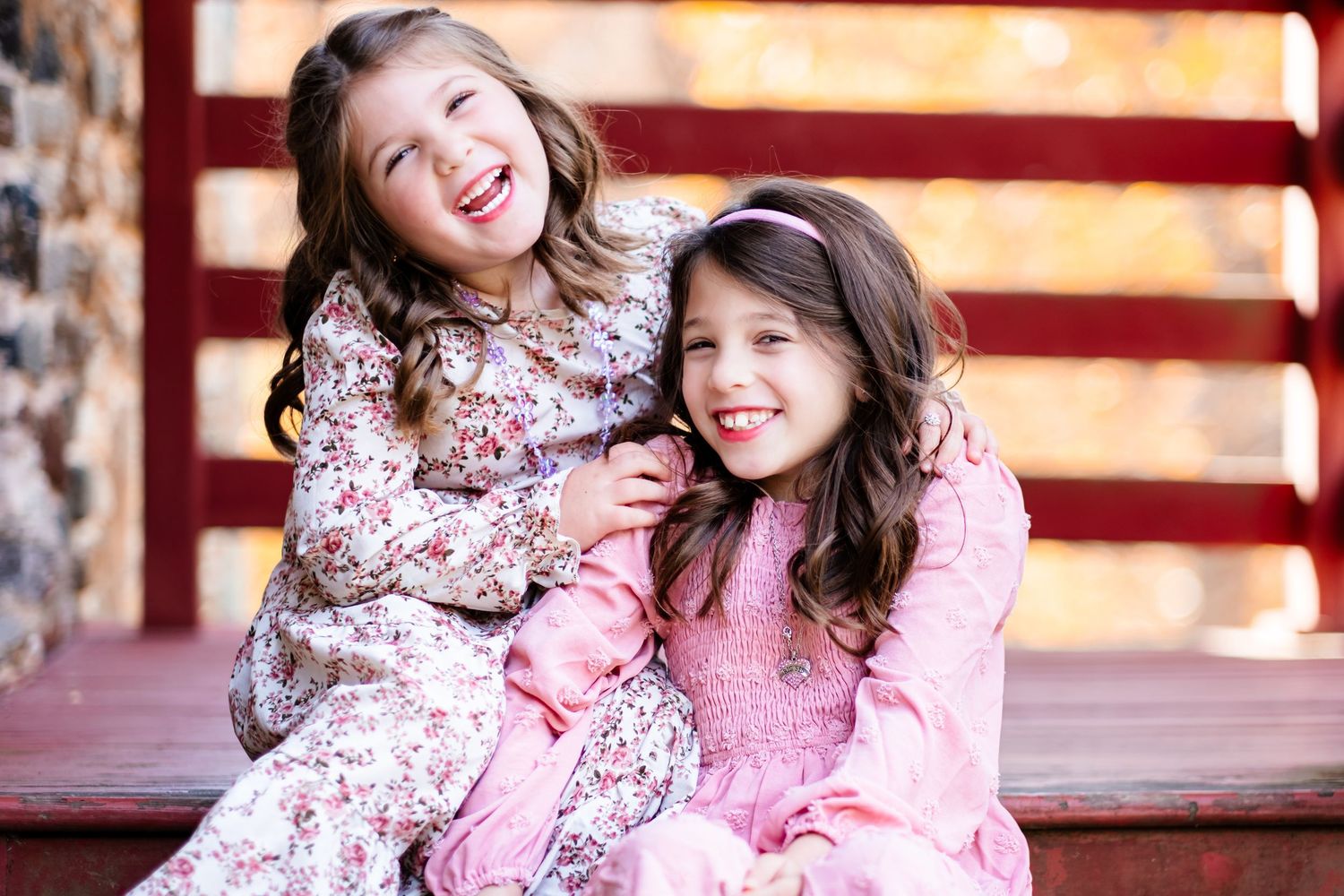 Two smiling girls in pink floral dresses pose together on wooden steps near a red fence in autumn.