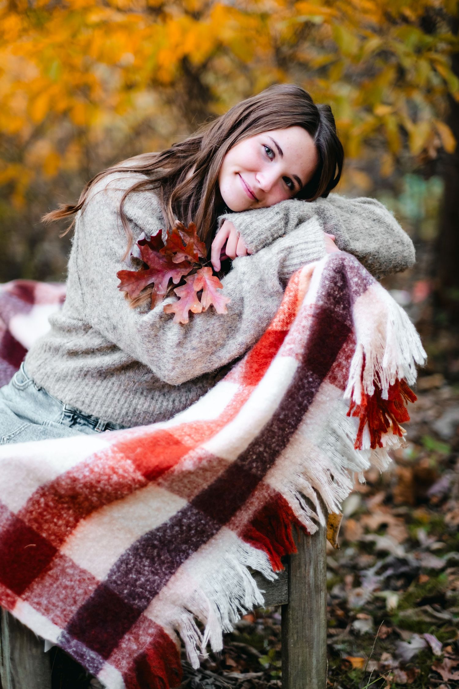 Young woman in cozy sweater holding autumn leaves, leaning on a plaid blanket in a colorful fall forest setting.