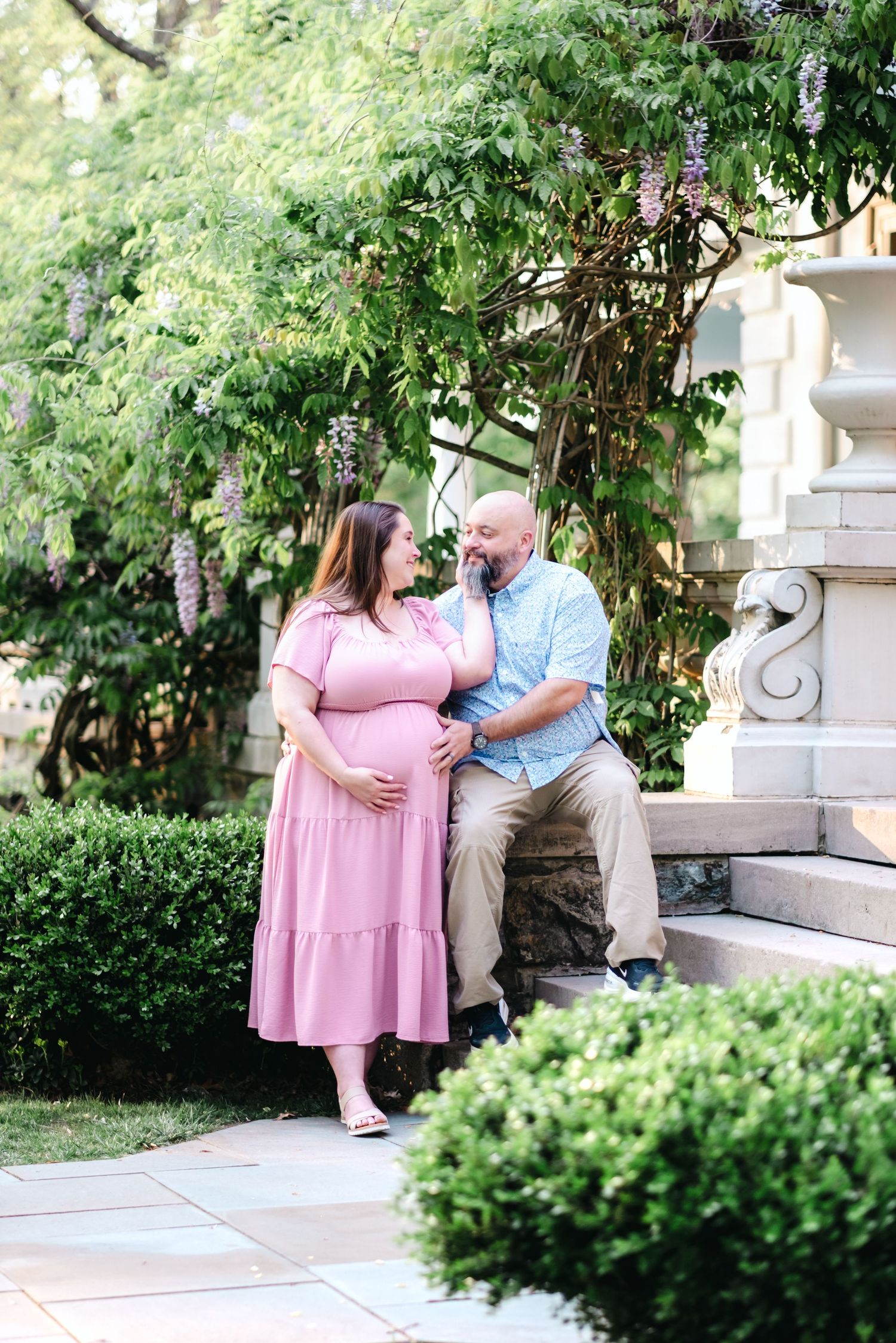 Pregnant woman in pink dress with partner in garden setting during maternity photo session.
