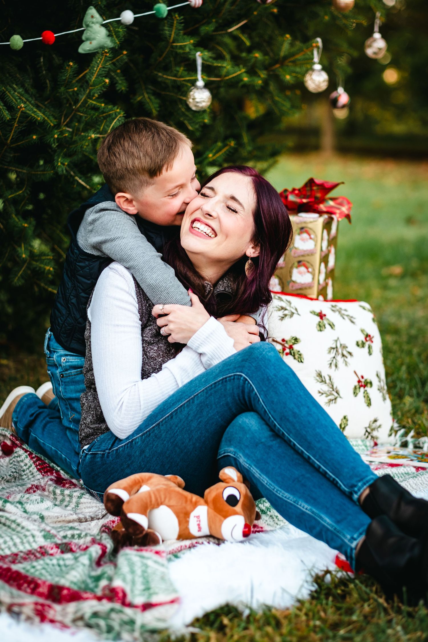 A joyful mother laughs as her young son hugs and kisses her cheek during a festive outdoor Christmas photo session.