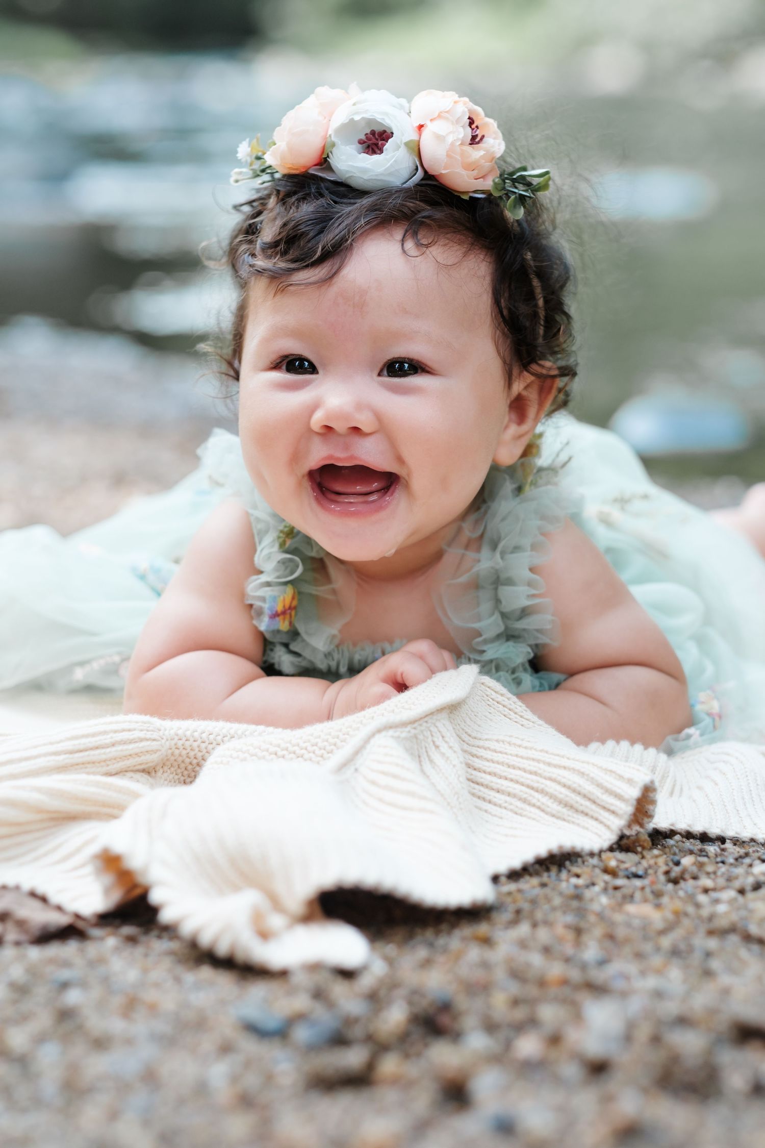 Smiling baby girl wearing a floral crown and mint dress, lying on a blanket near a serene outdoor setting.