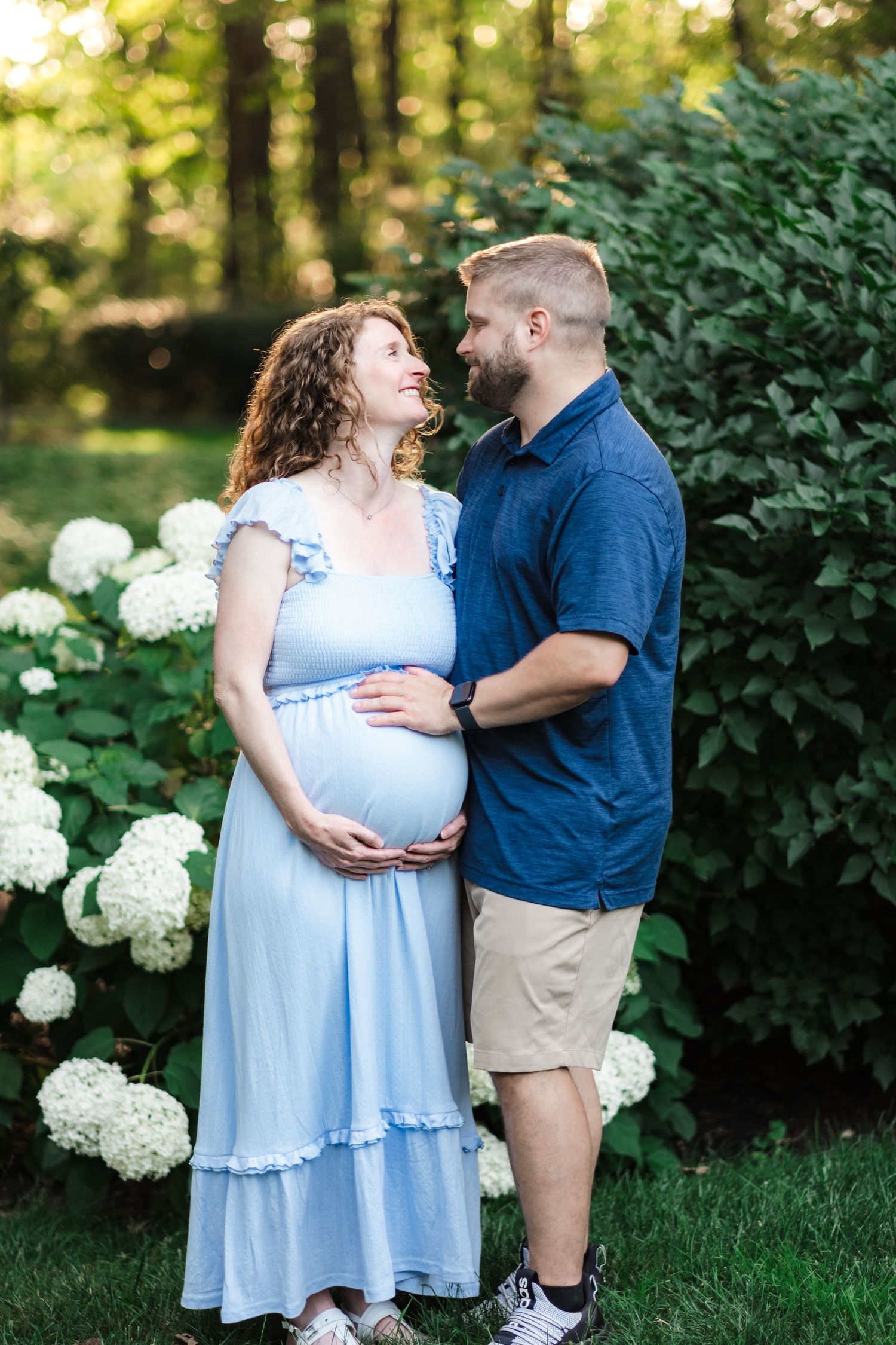 Pregnant woman in blue dress and man in navy shirt share a tender moment outdoors among white hydrangeas.