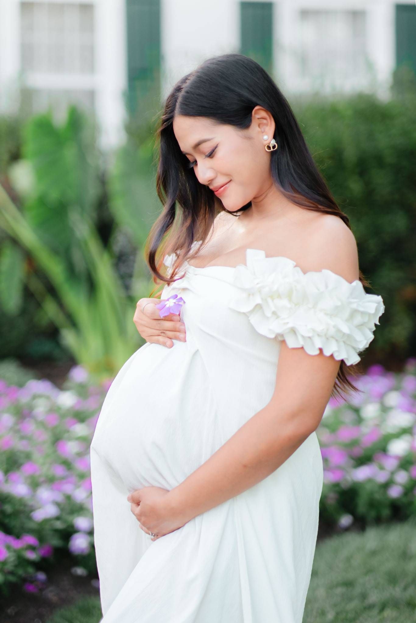 Pregnant woman in white off-shoulder dress holding flowers, smiling at baby bump in a garden.