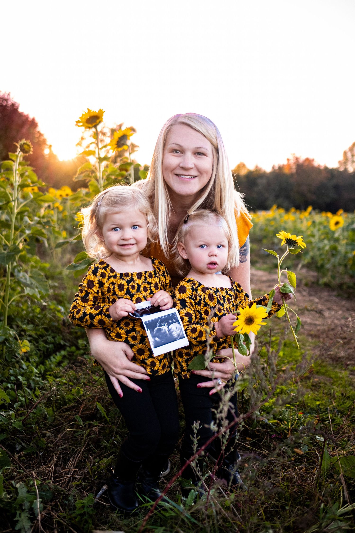 A mother and two young girls in matching sunflower outfits pose in a sunflower field holding an ultrasound photo.