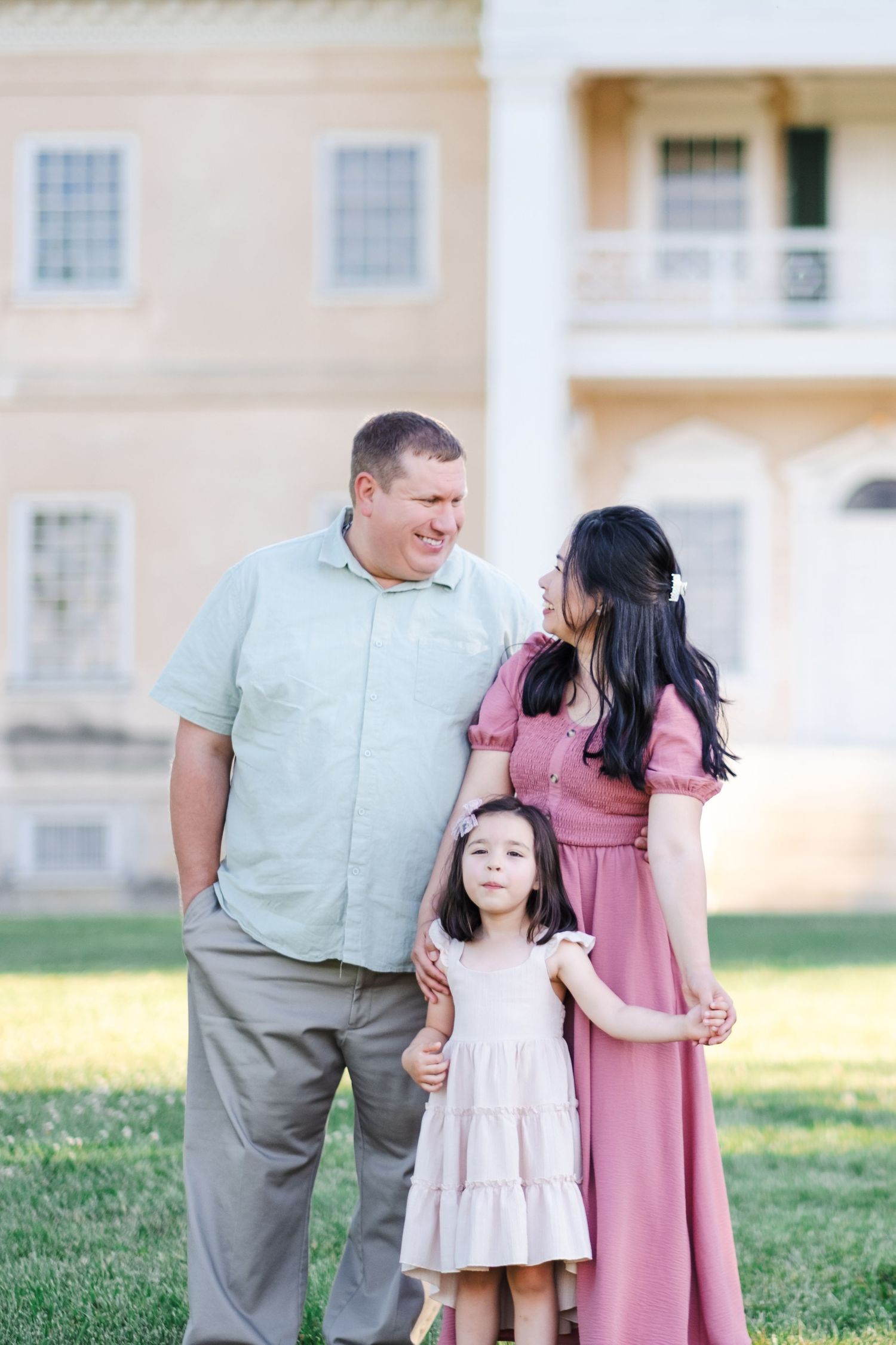 Family of three posing outdoors in front of a grand white colonial mansion on a sunny day.
