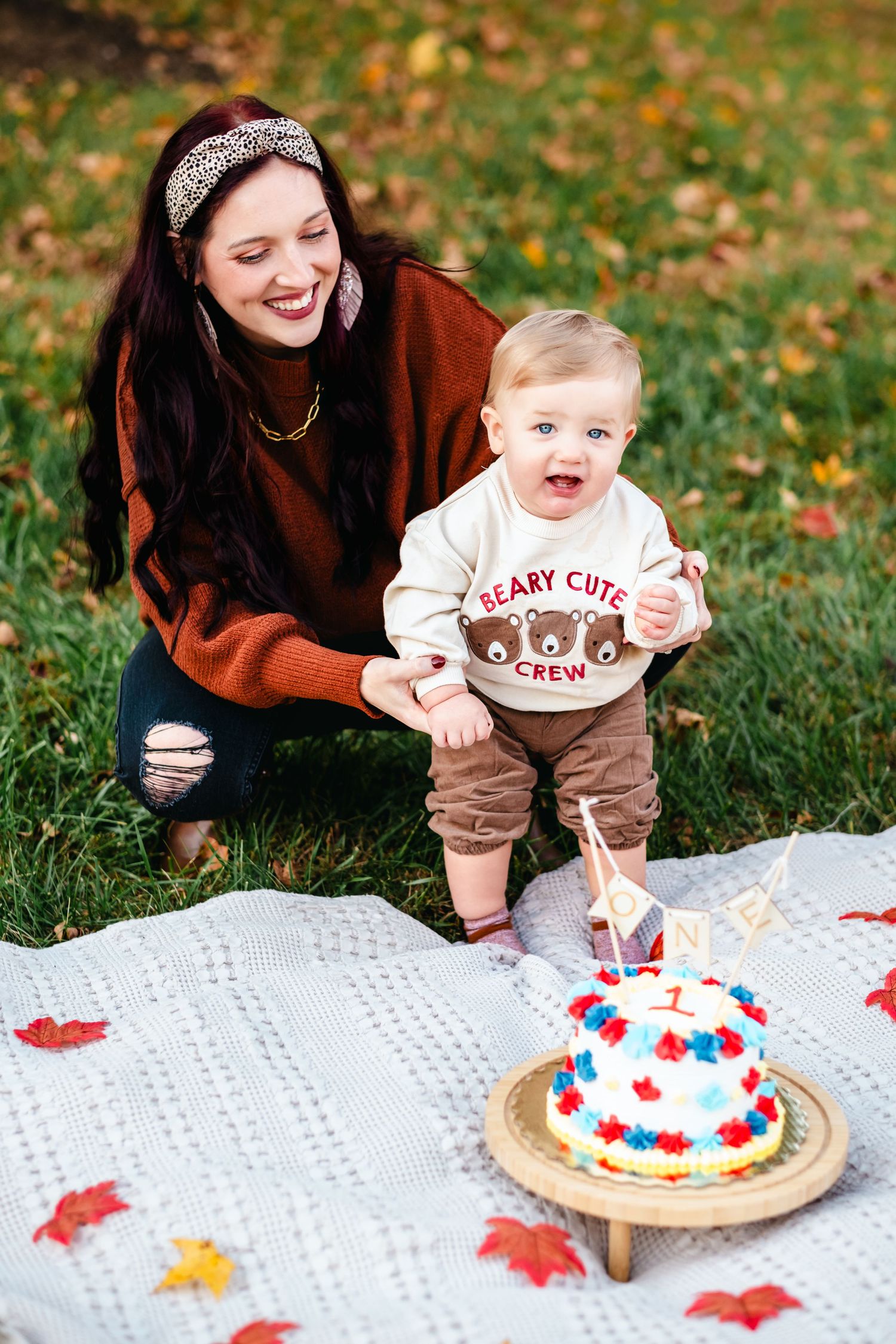 A smiling mother kneels beside her baby boy in a bear crew sweater at an outdoor first birthday cake smash session.