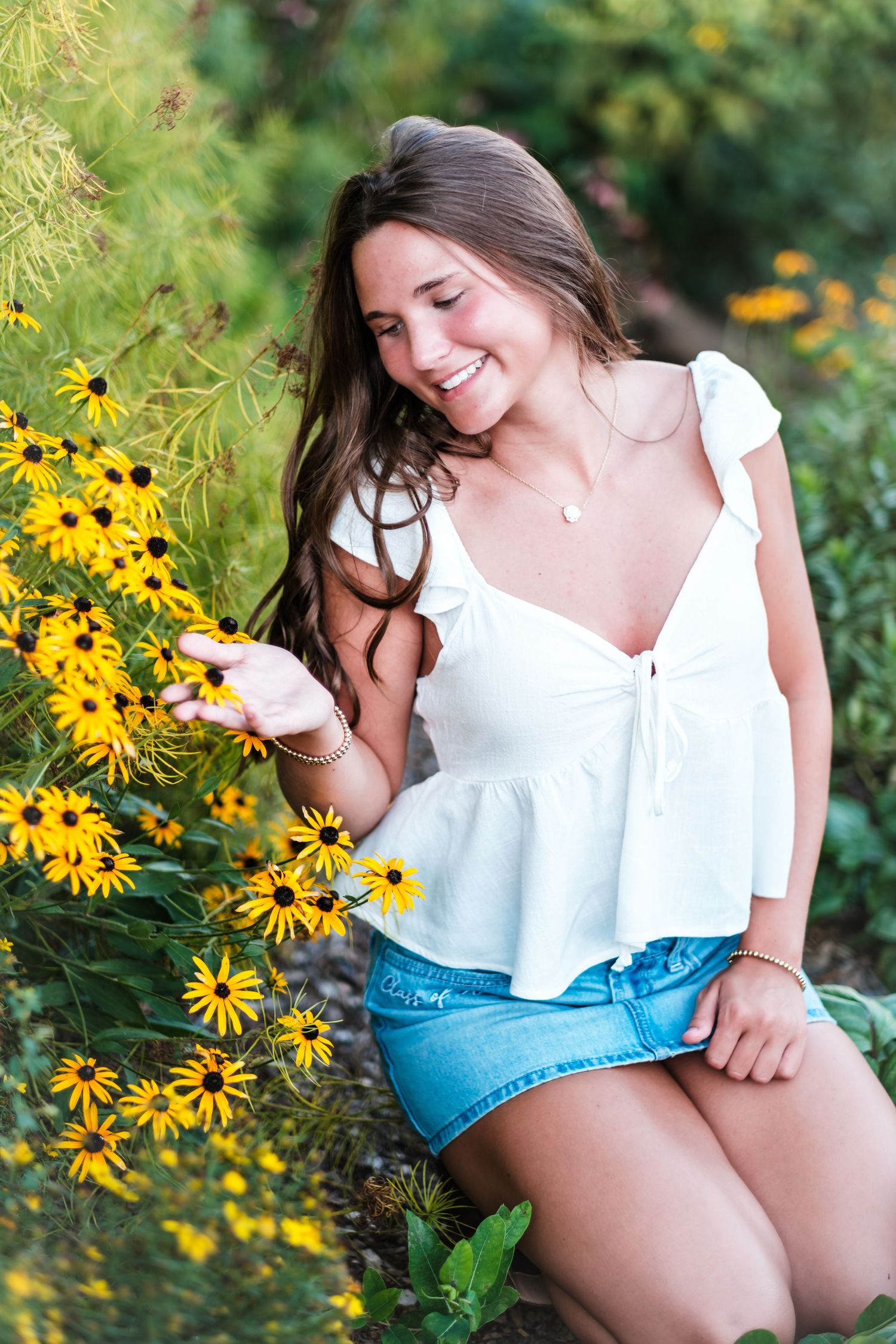 Young woman in white top and denim skirt smiling while touching yellow black-eyed Susan flowers in a lush garden.