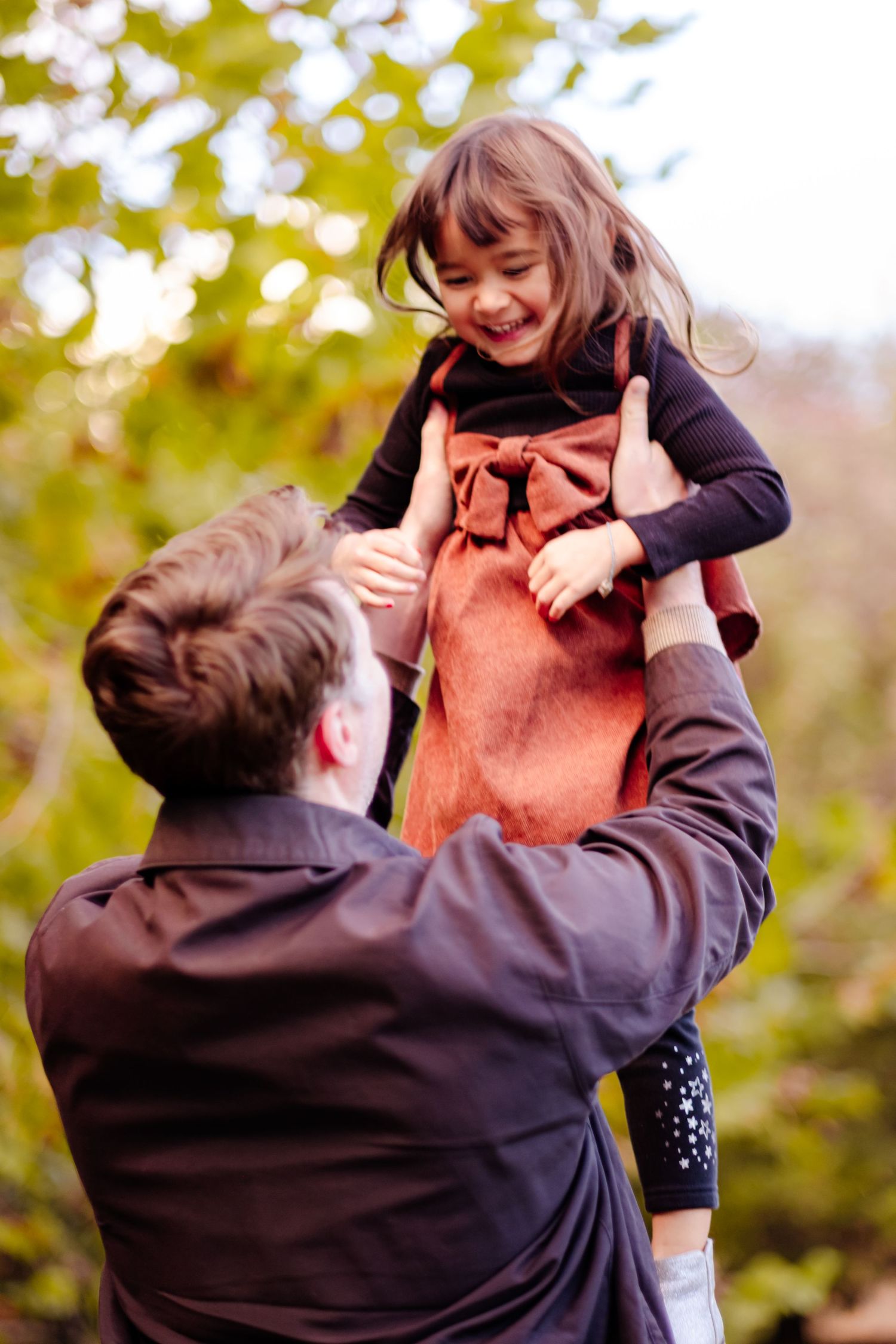 Father lifting smiling young girl in orange dress outdoors in autumn park, sharing a joyful moment together.