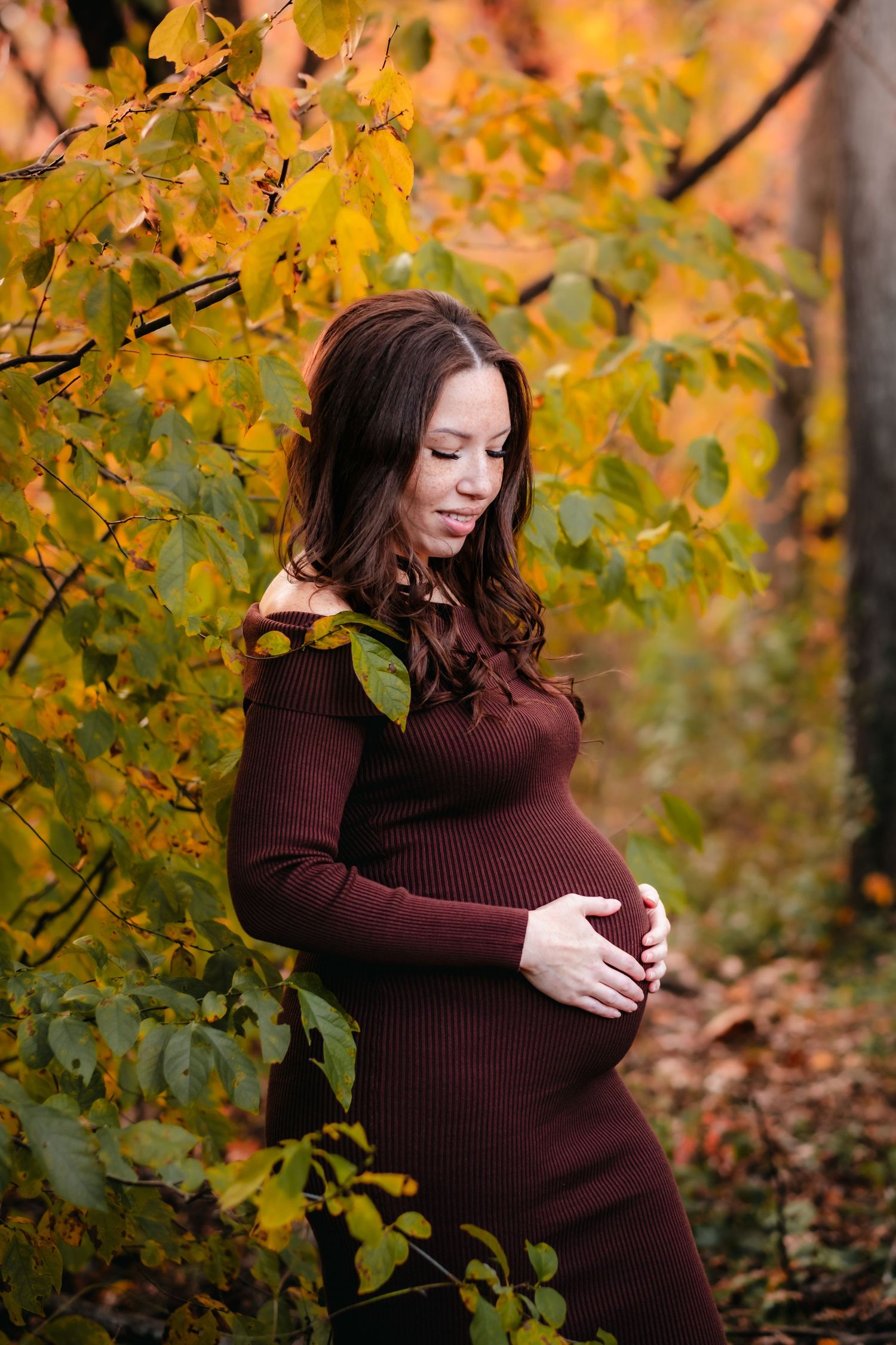 Pregnant woman in burgundy dress cradling baby bump among vibrant autumn foliage in a serene outdoor setting.