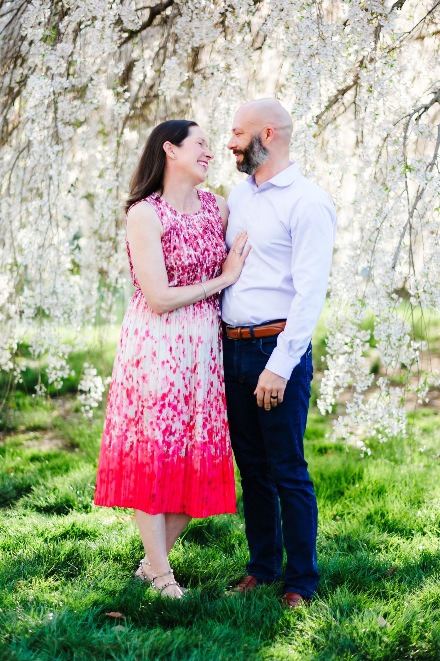 A couple smiles at each other under blooming white cherry blossom trees, woman in pink floral dress, man in white shirt.