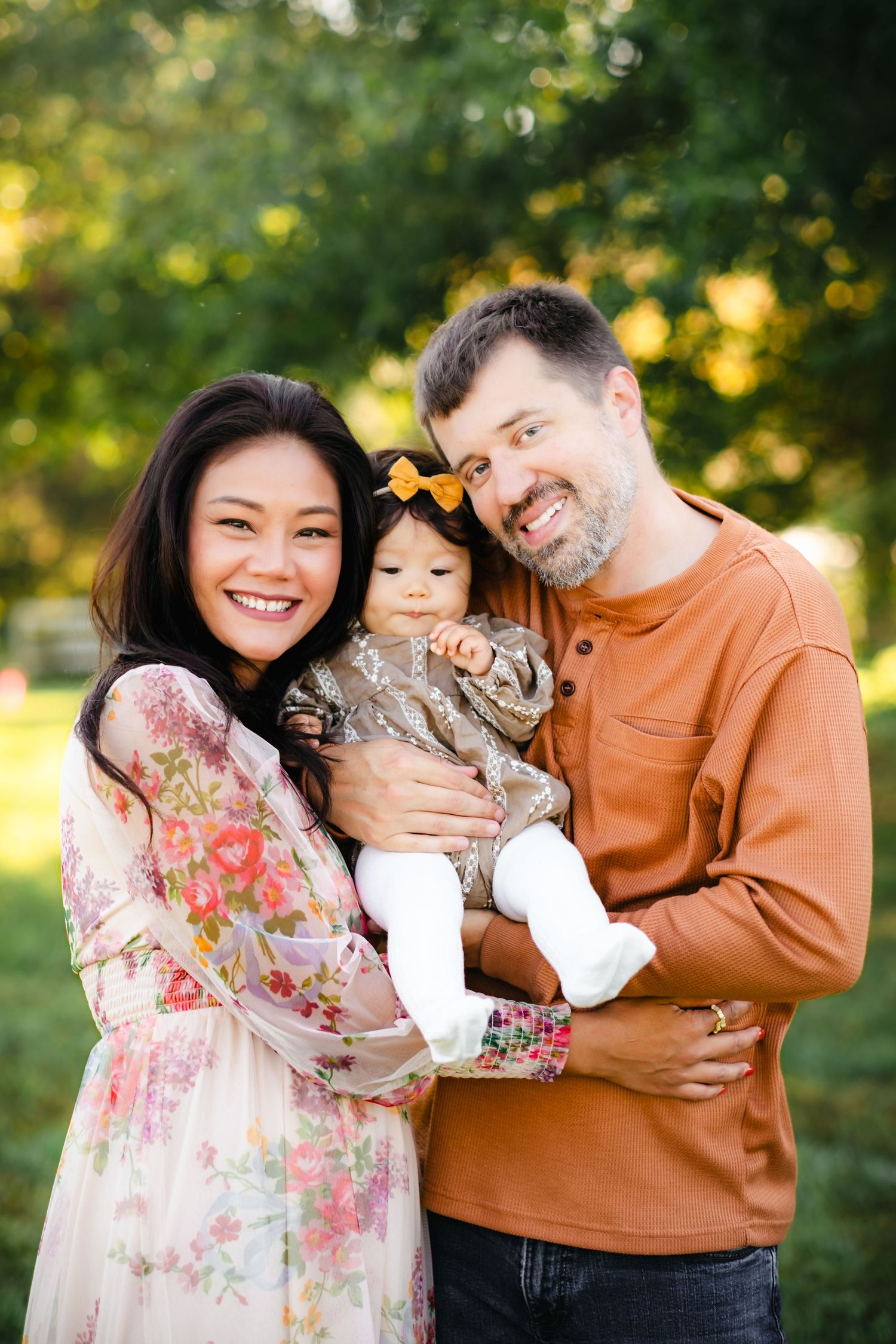 Smiling family portrait with a woman in floral dress, man in rust shirt, and baby girl in a park setting.