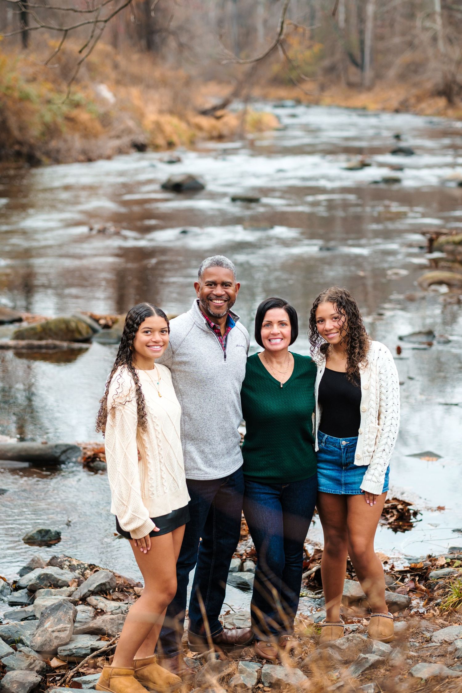 Family of four smiling together by a rocky stream in autumn woods, wearing casual fall outfits.