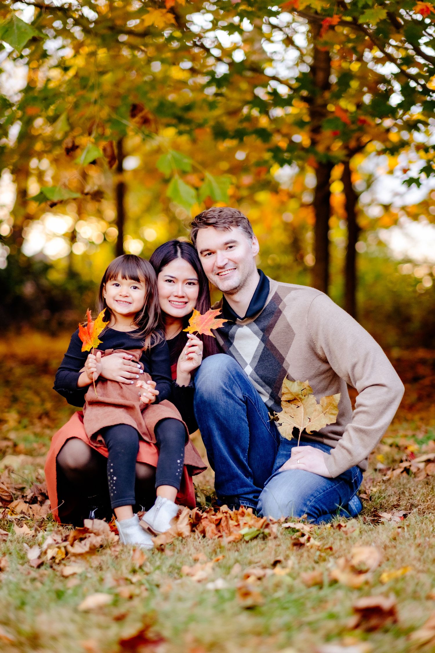 Family of three posing with colorful autumn leaves in a park, smiling warmly during a fall photo session.