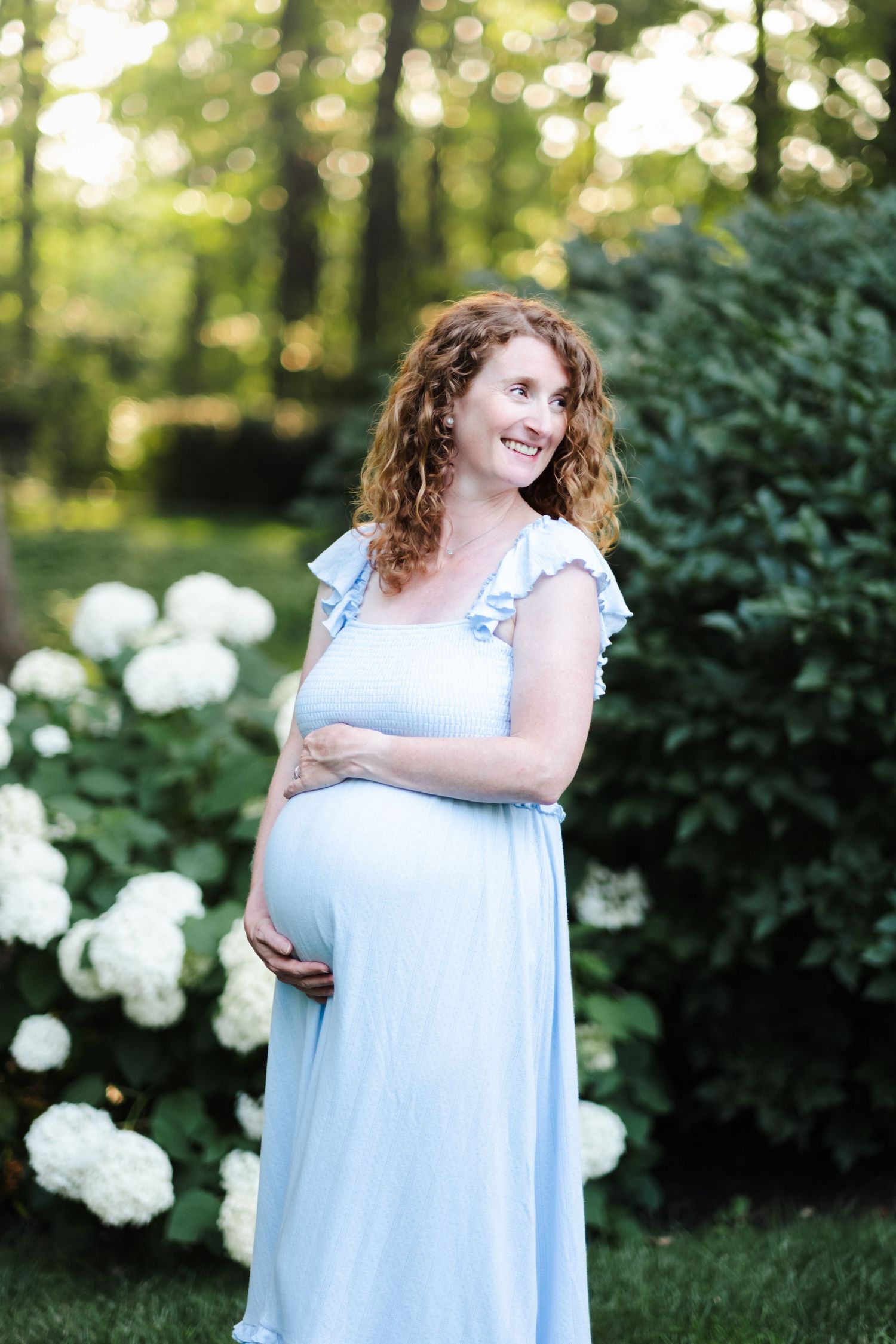 Smiling pregnant woman in light blue dress holding baby bump outdoors near white hydrangeas and green foliage.