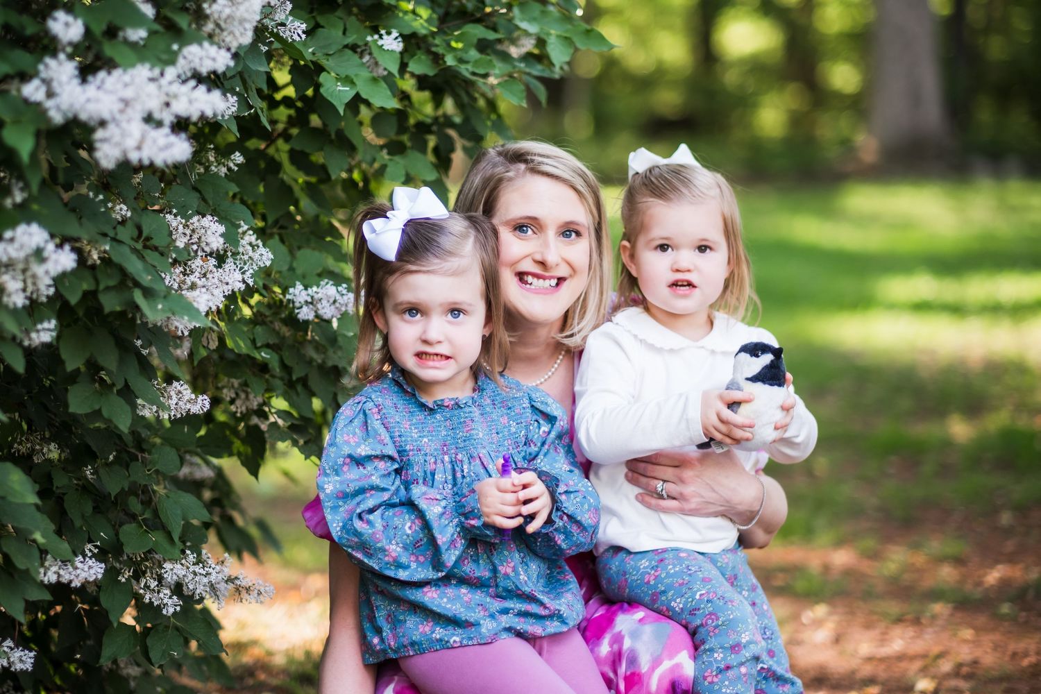 A family sits together outdoors near flowering bushes in a sunlit garden setting on a warm day.