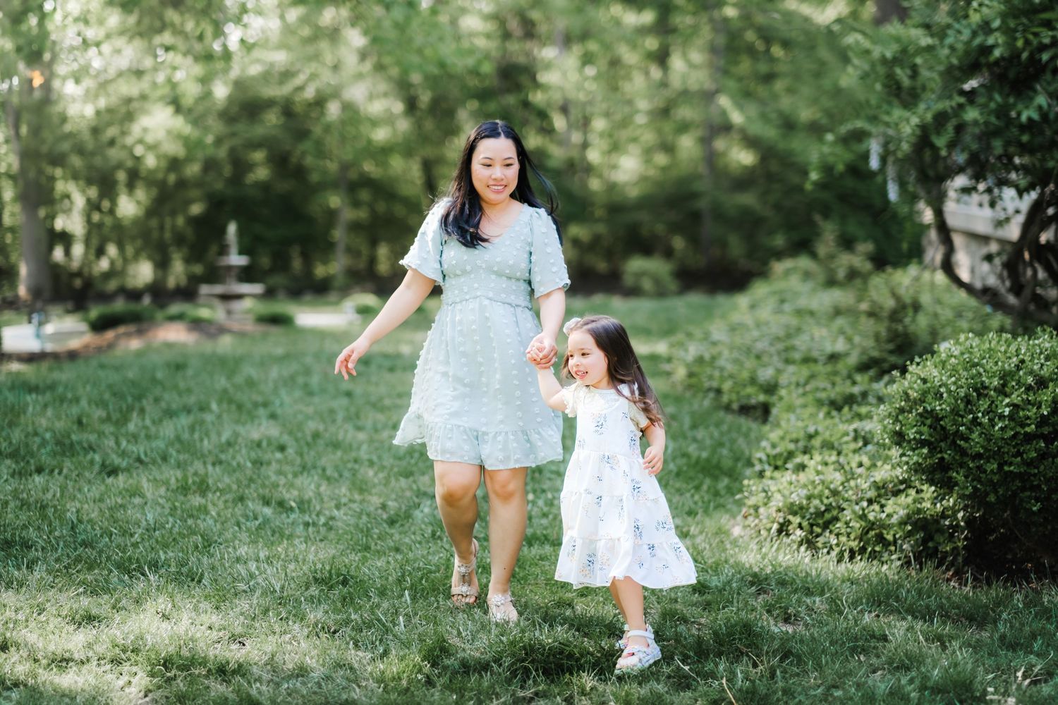 A parent and child in light blue and white dresses walk together through a lush green park on a sunny day.