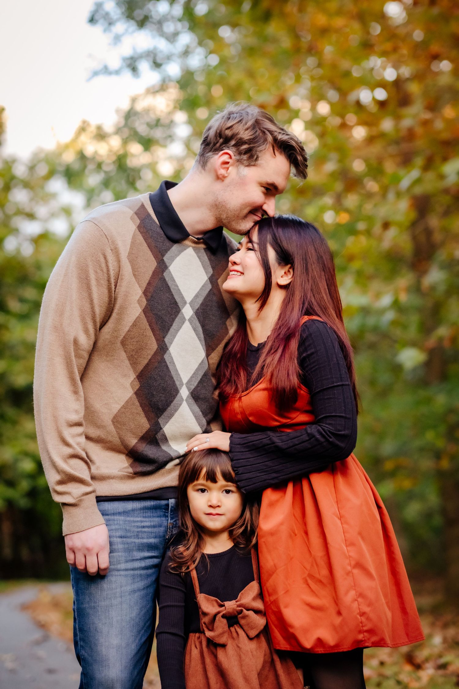 Family of three posing outdoors in autumn, parents sharing a tender moment while young daughter stands between them.