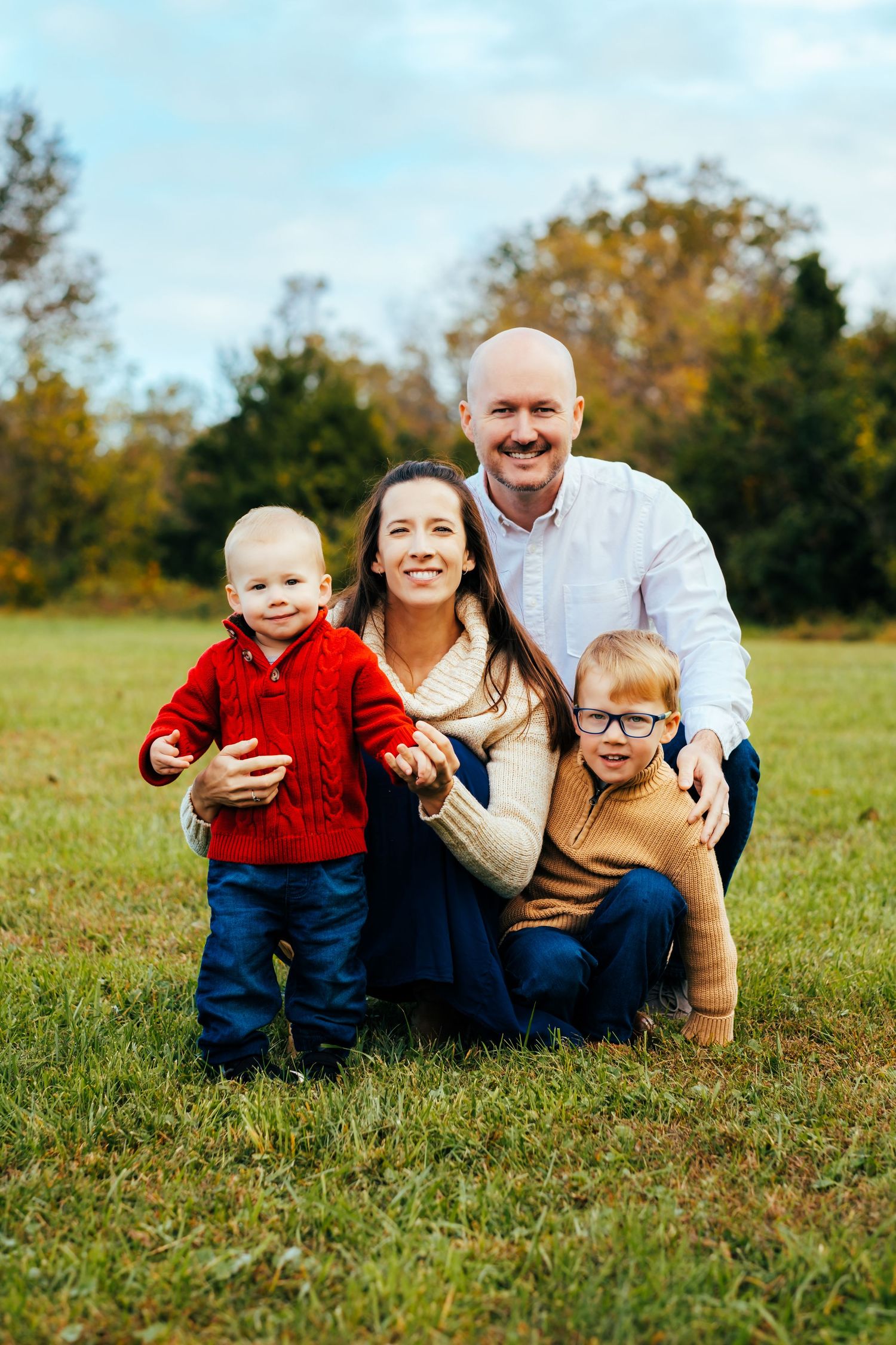 Family of four posing outdoors in autumn, parents with two young boys wearing colorful sweaters on green grass with fall foliage.