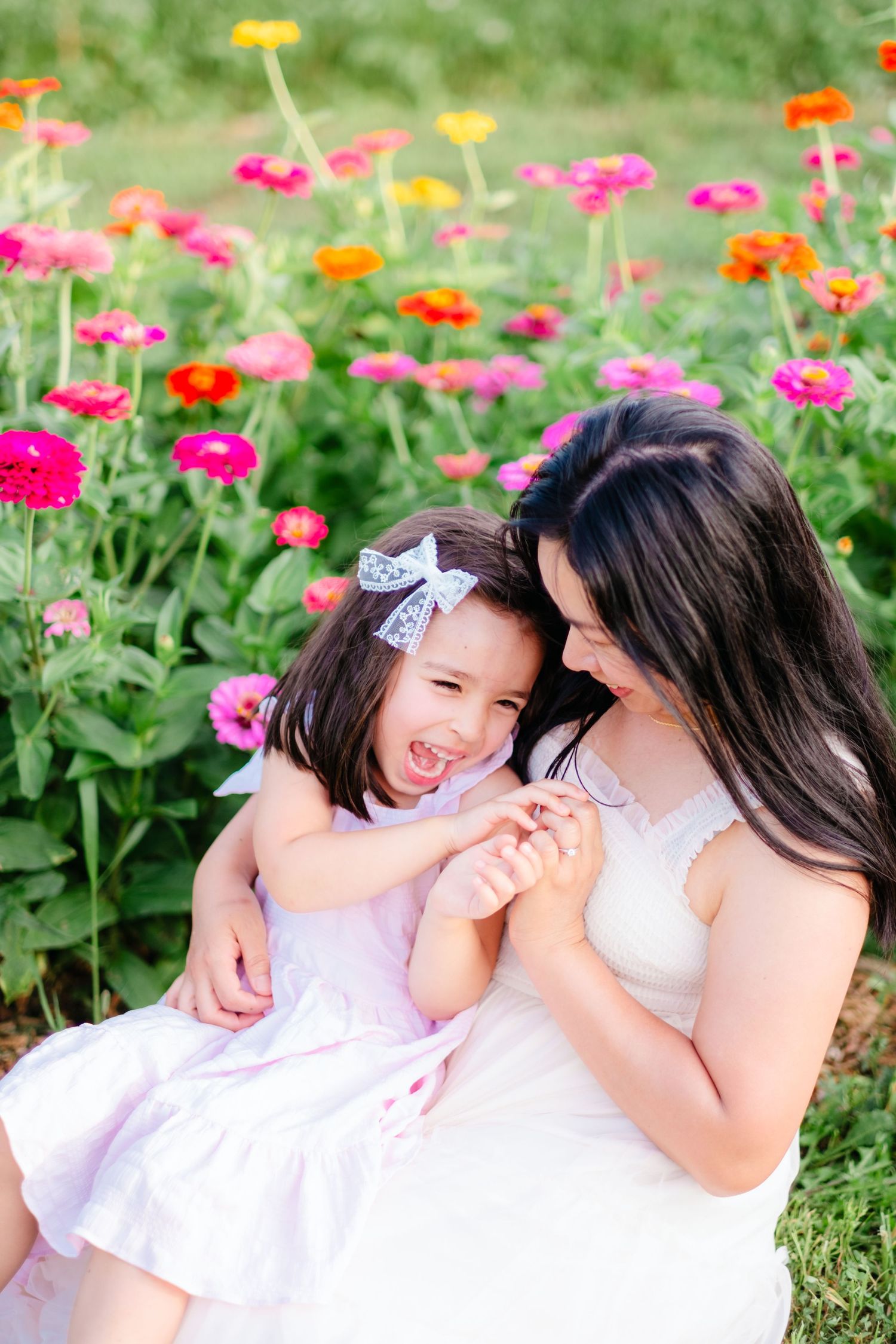 Mother and daughter laughing together in a colorful zinnia flower garden, girl wearing pink dress with bow.