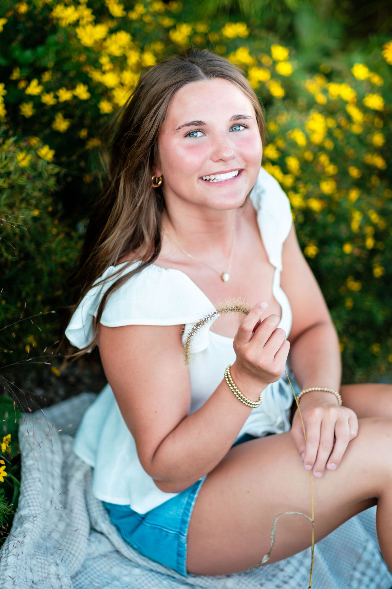 Smiling teenage girl in white top and denim shorts sitting outdoors among vibrant yellow flowers.