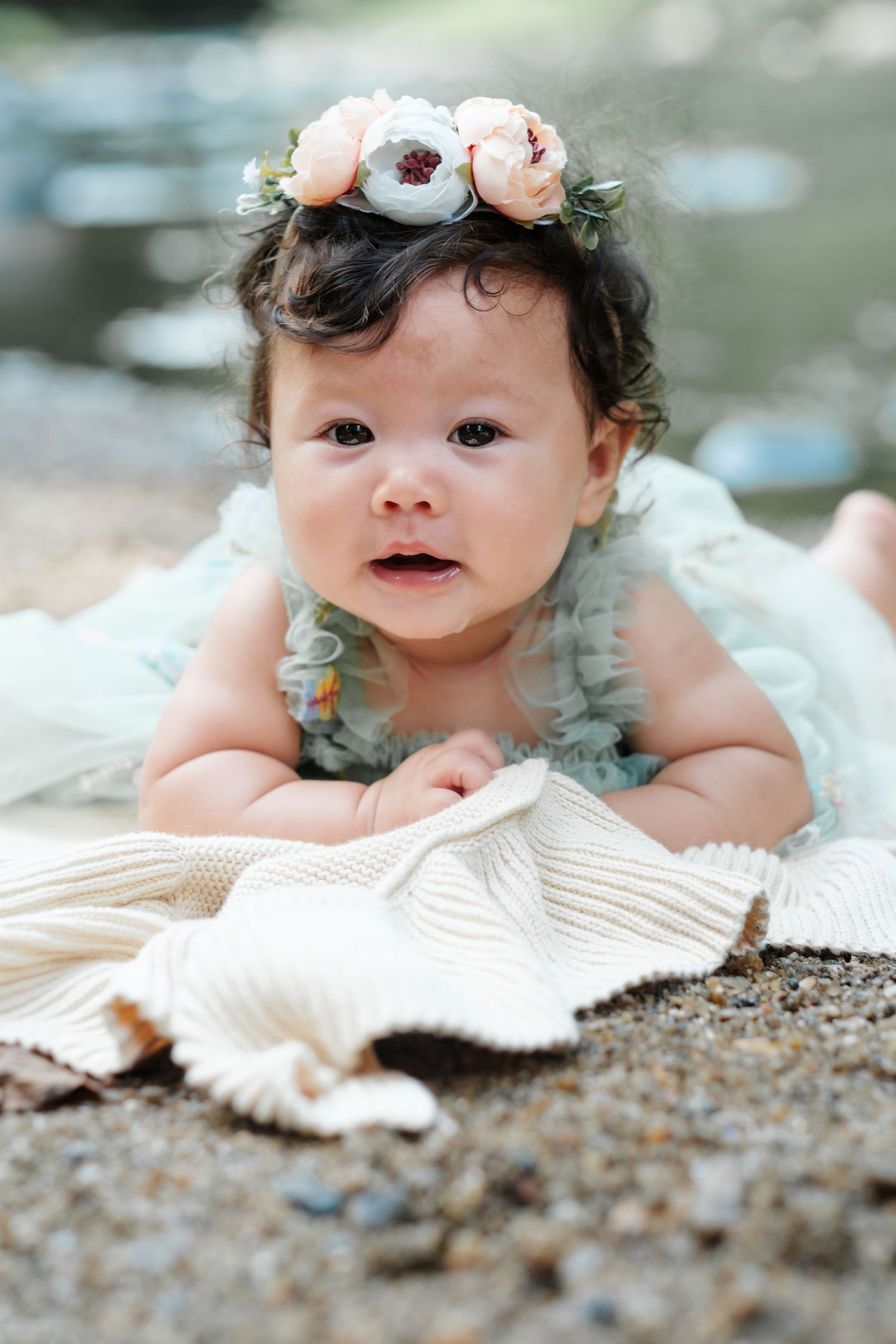 Baby girl in floral crown and blue ruffle dress lying on knit blanket outdoors near water.