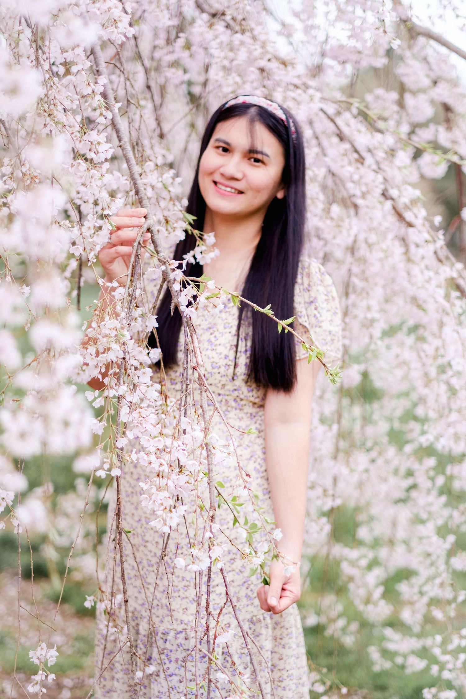 A woman in a floral dress poses among blooming cherry blossom trees during spring.