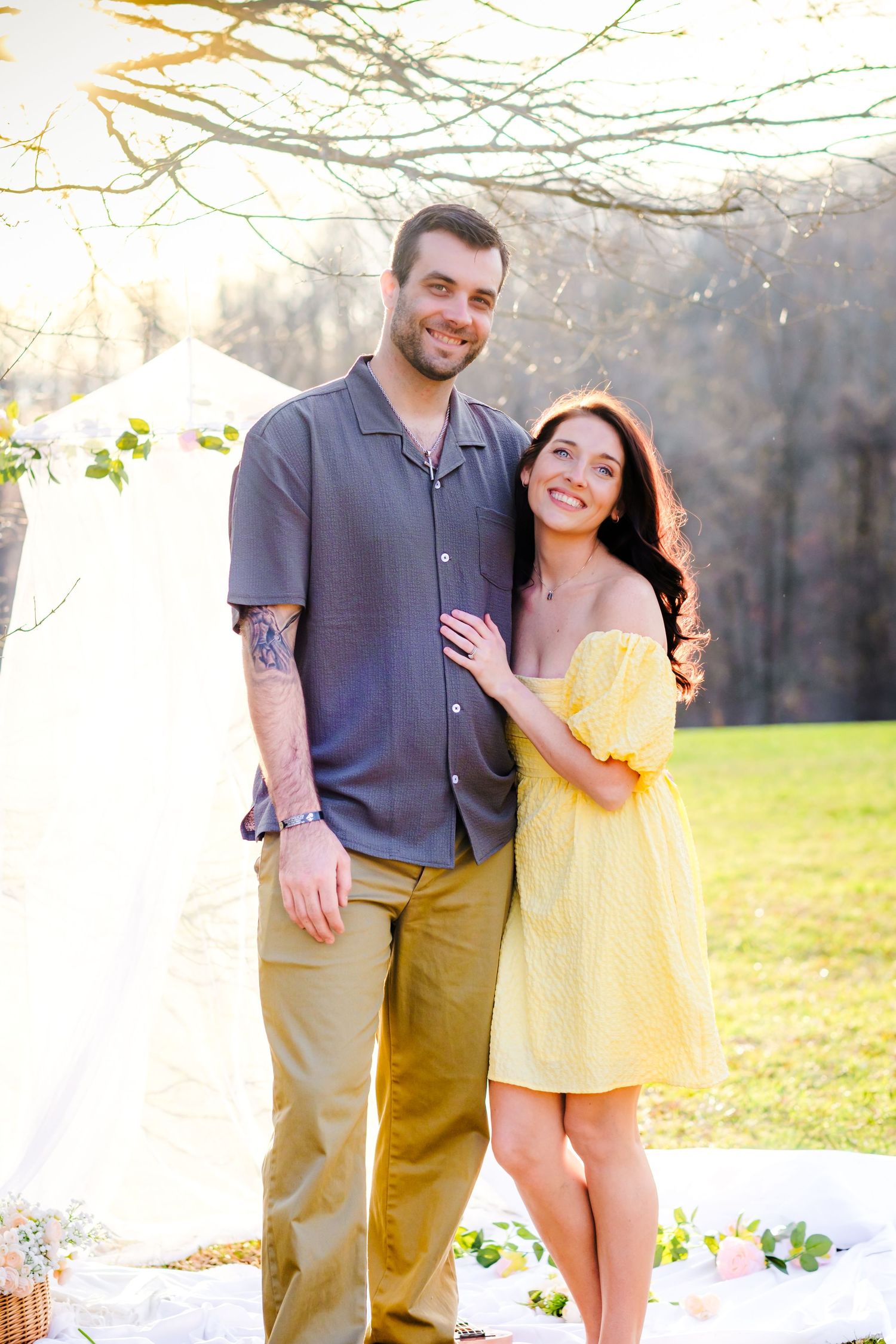 A smiling couple poses outdoors, man in gray shirt and khakis, woman in yellow off-shoulder dress, with greenery behind.