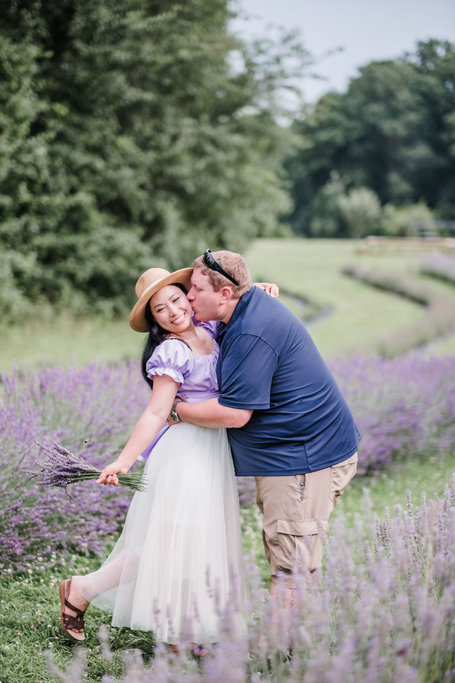 Couple sharing a kiss in a lavender field, woman in purple dress and straw hat holding fresh lavender bouquet.