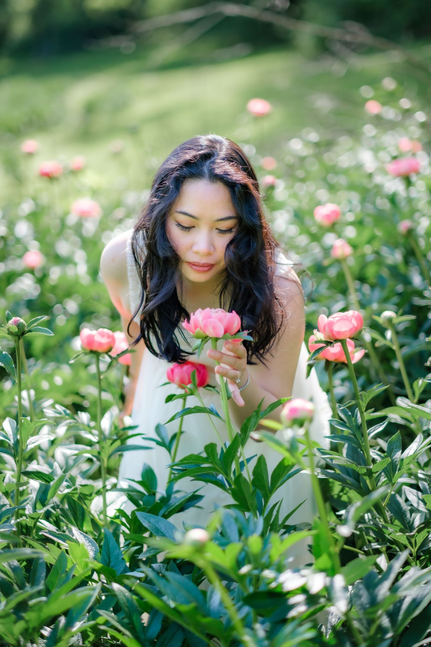 Young woman in white dress smelling pink peonies in a lush green garden field.