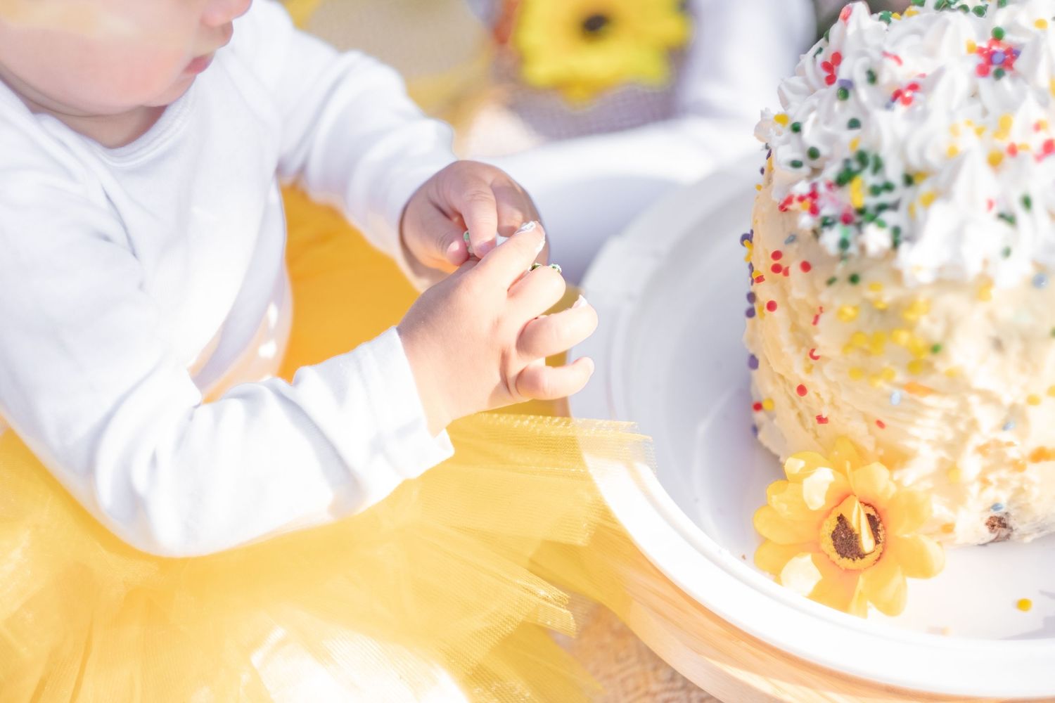 Baby in yellow tutu reaching into white sprinkle birthday cake decorated with sunflowers.
