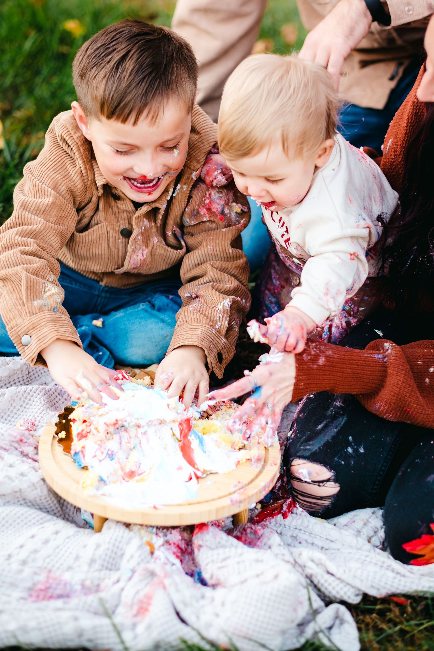 Two young children joyfully smashing a colorful birthday cake together outdoors on a white blanket.