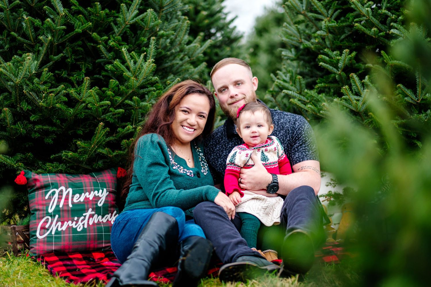 Family Christmas photo at a tree farm with parents and baby on a red plaid blanket with Merry Christmas sign.