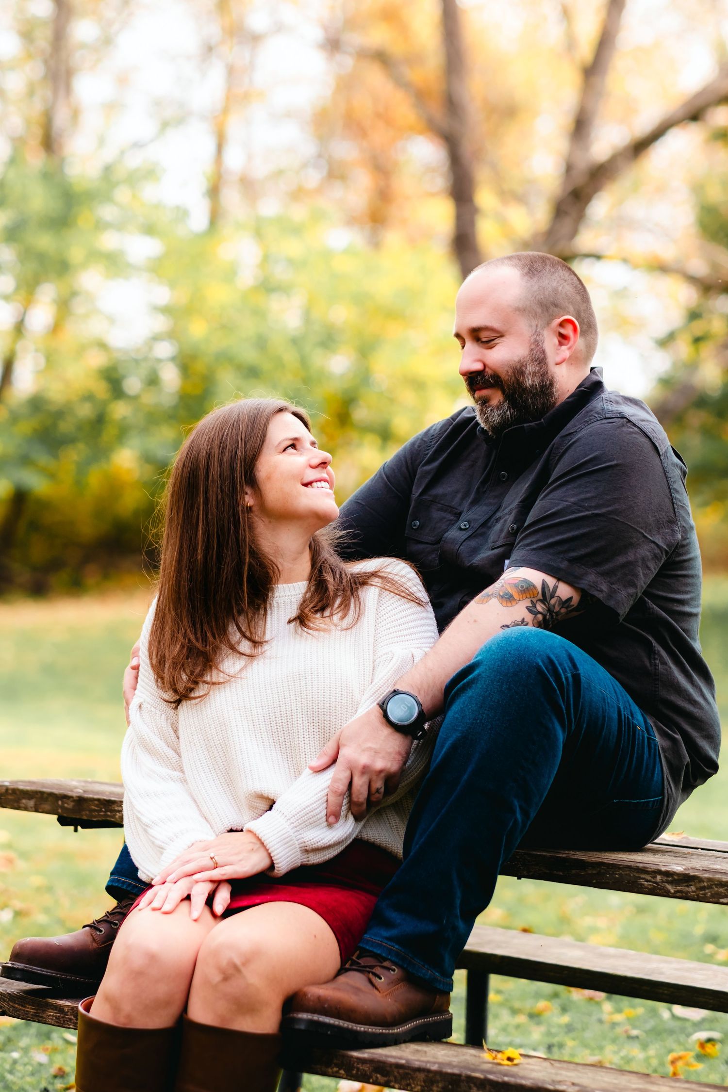 A couple sits on a park bench surrounded by autumn foliage, sharing a warm and loving glance at each other.