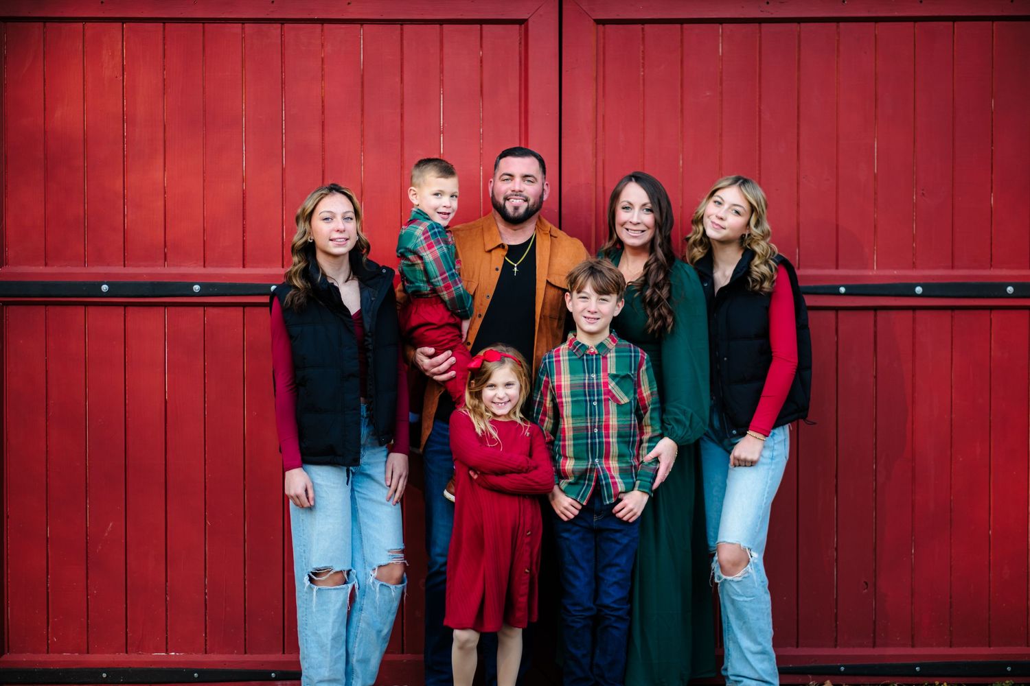Family of seven posing in front of red barn doors for a festive holiday portrait.