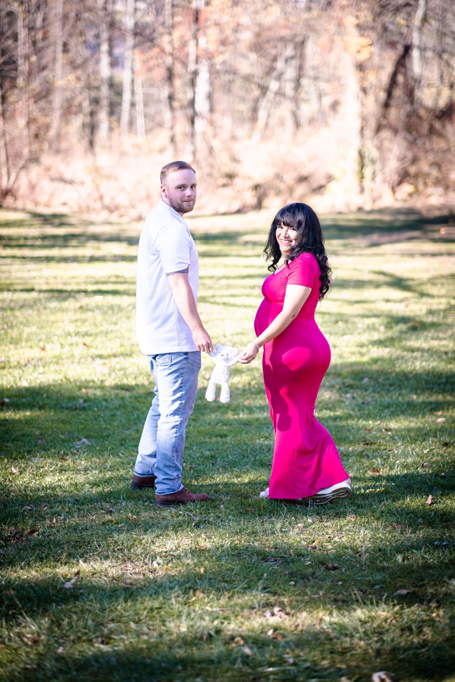 Pregnant woman in pink dress holds hands with man holding teddy bear outdoors in park during fall season.
