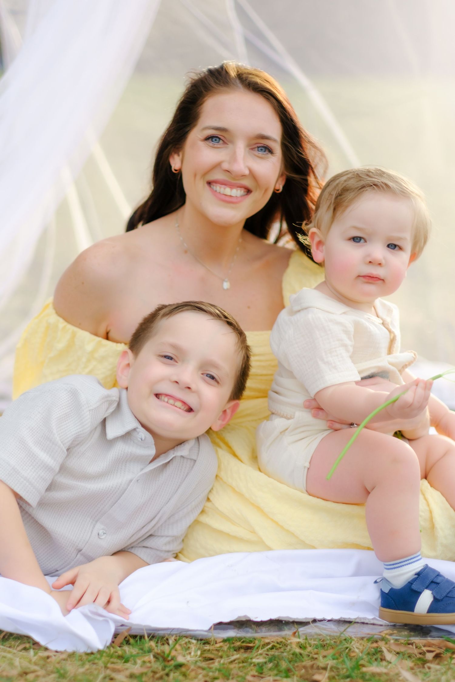 Cheerful mother in yellow dress smiles with two young boys on white blanket during outdoor family session.