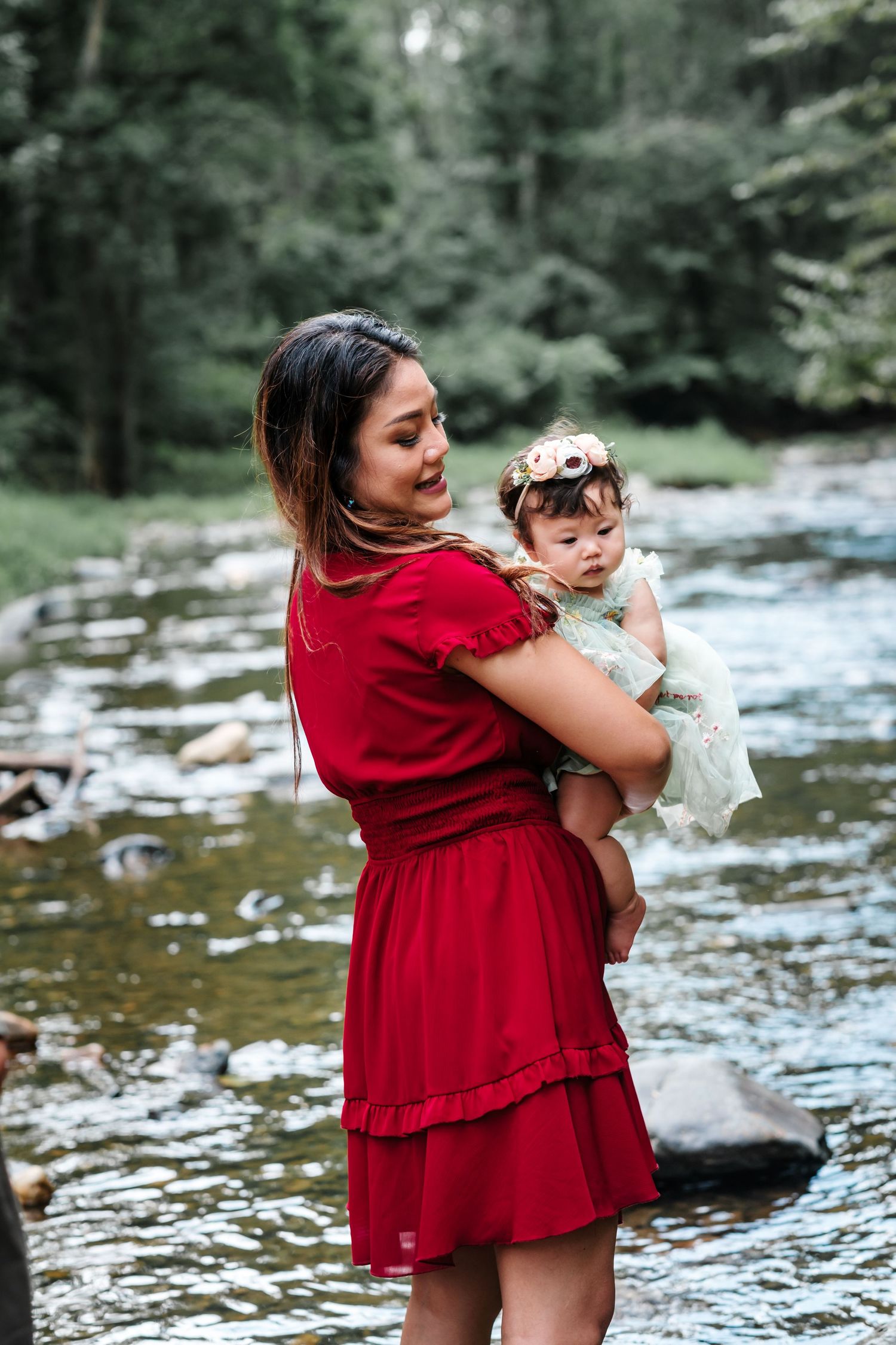 A mother in a red dress holds her baby girl wearing a floral crown near a rocky stream in a lush forest.