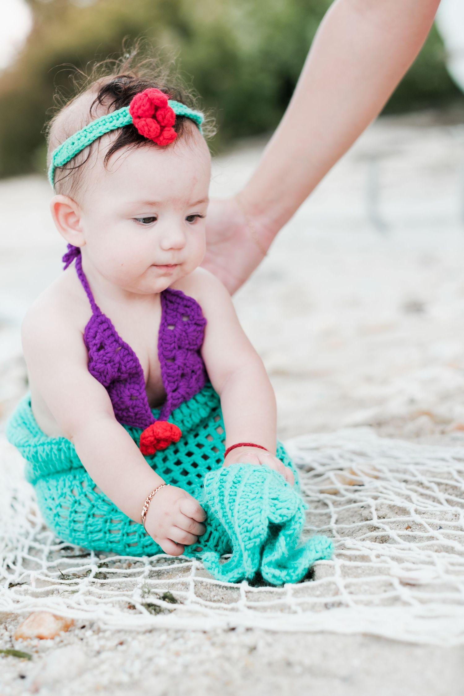 Baby girl in crochet mermaid costume with teal skirt and purple top sits on fishing net at beach.