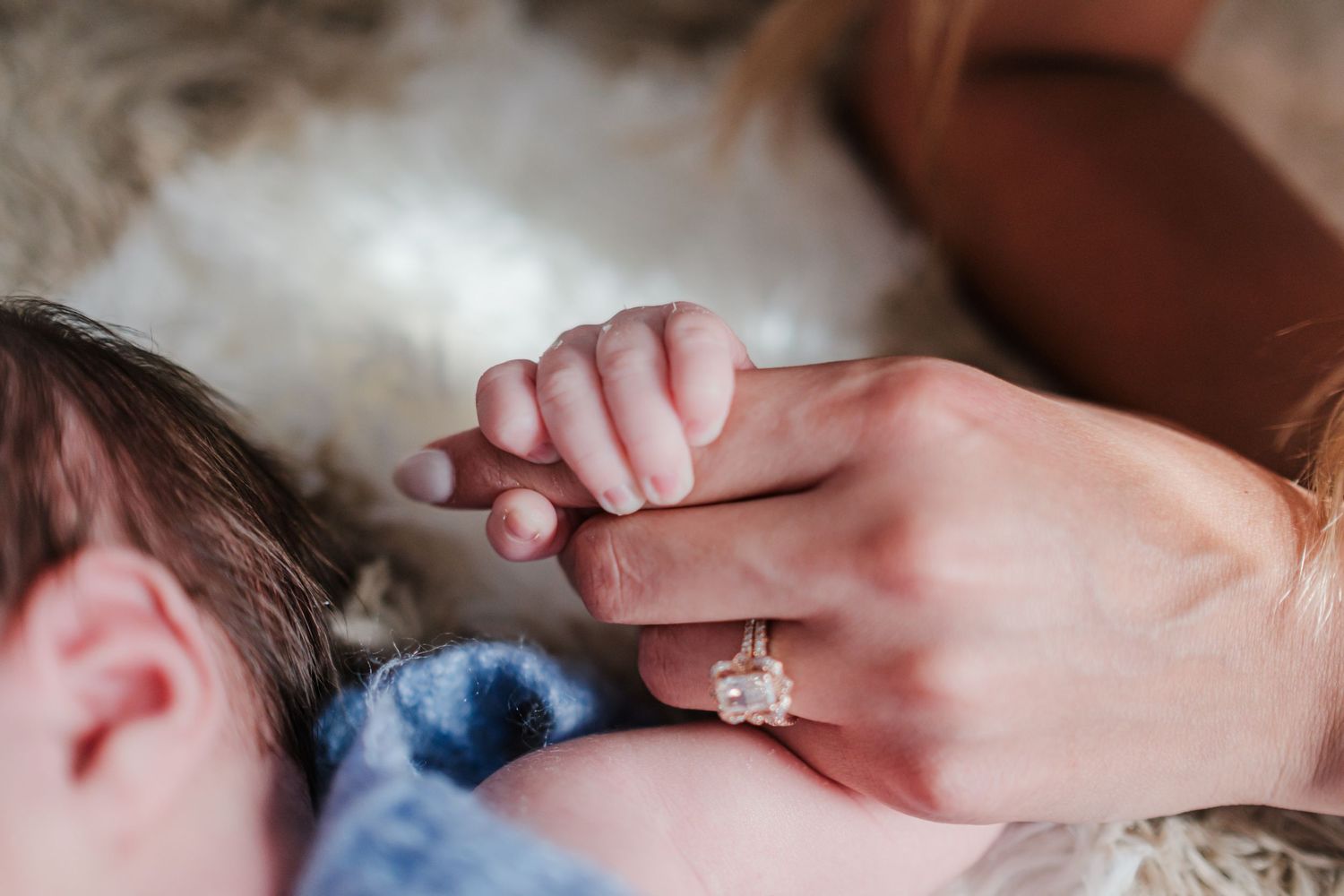 A newborn baby grasping an adult's finger wearing a diamond engagement ring, capturing a tender mother-child bonding moment.