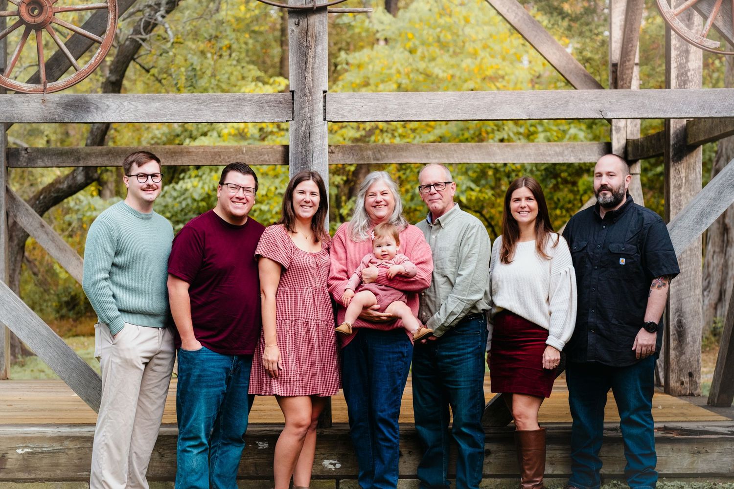 Family of seven posing outdoors near rustic wooden gate, smiling together while holding a newborn baby in autumn setting.