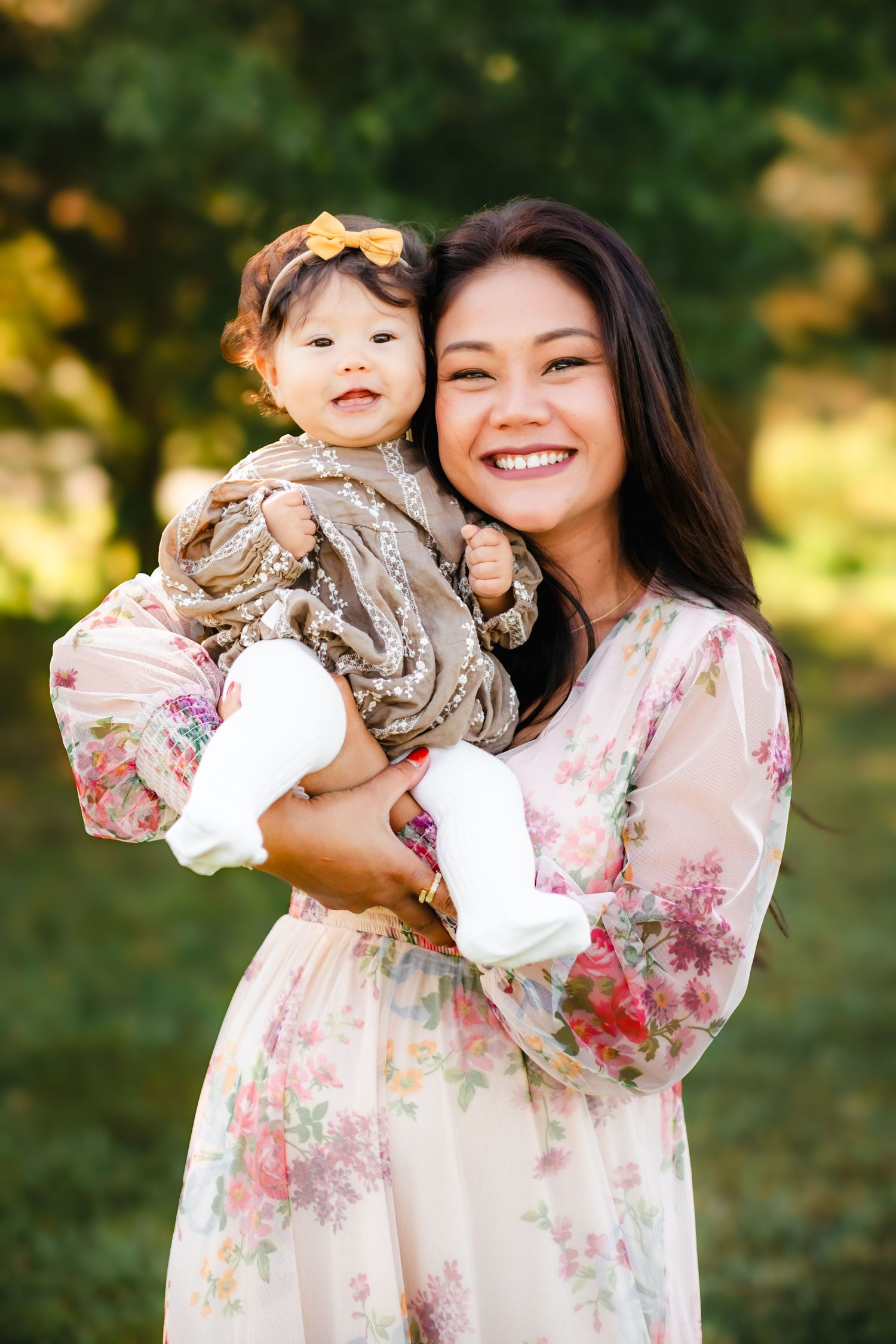 Smiling Asian mother in floral dress holding her baby girl outdoors in a lush green park setting.