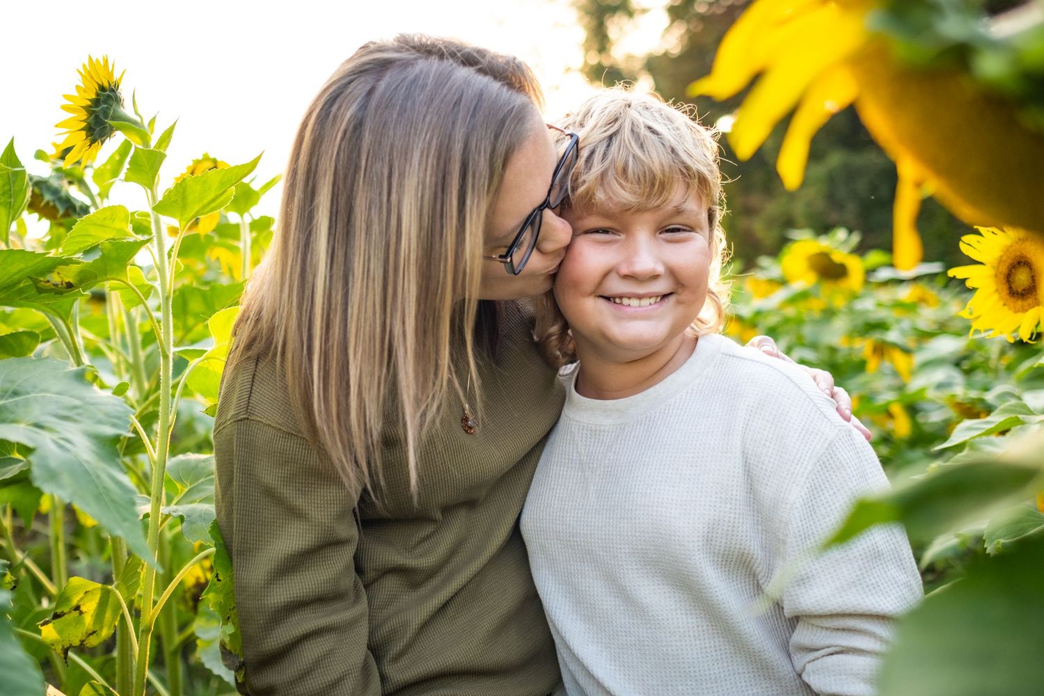 A mother kisses her smiling son surrounded by bright sunflowers in an outdoor sunflower field.