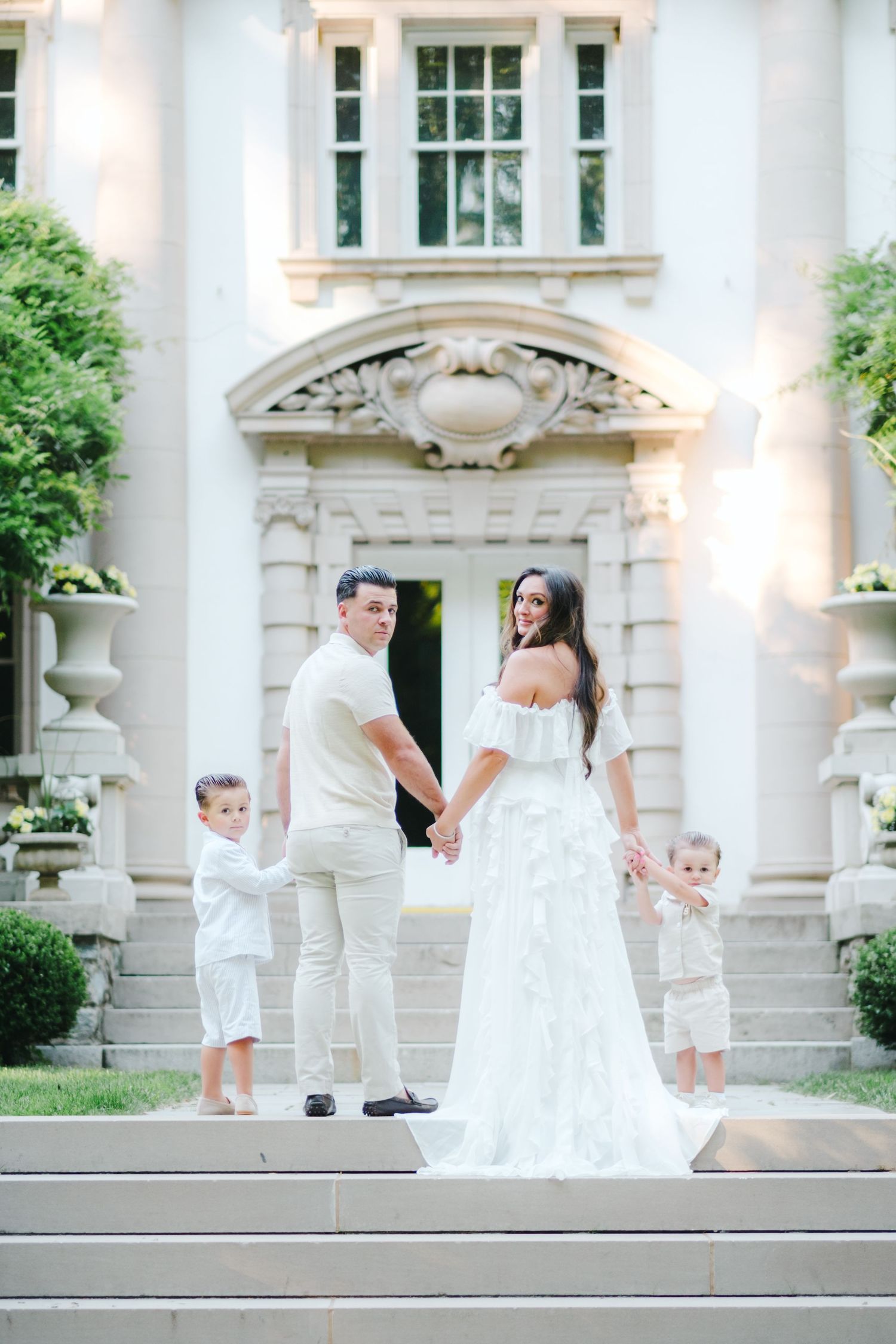 Family of four in white outfits posing on steps before an elegant mansion, parents holding hands with two young children.