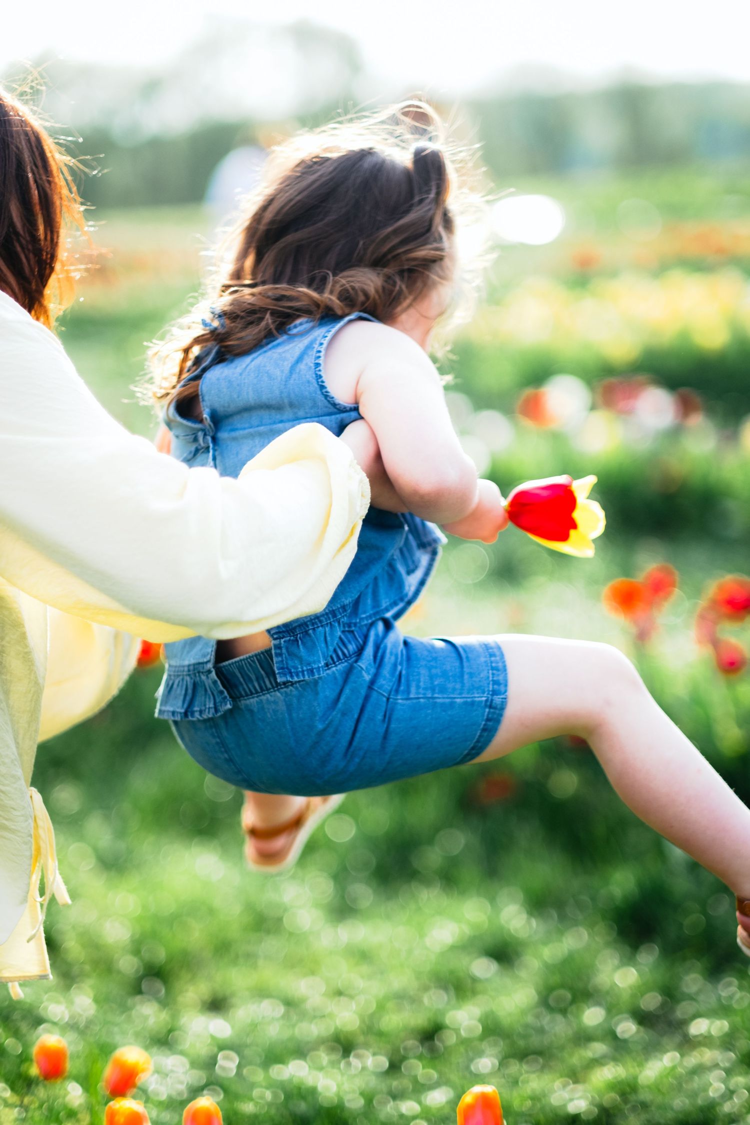 A mother swings her young daughter in a colorful tulip garden, the girl holding a red tulip while wearing a denim romper.