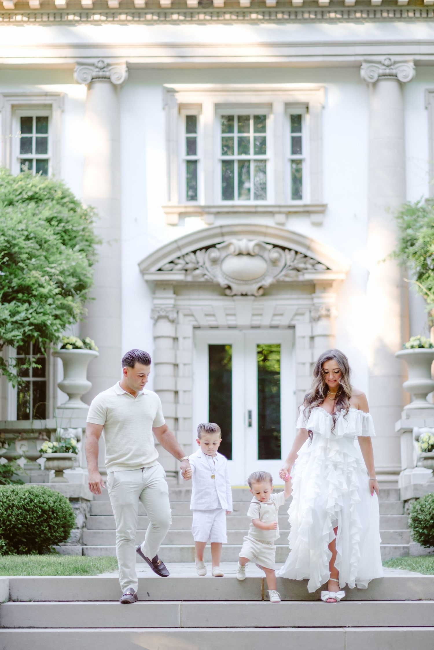 Family of four in white and cream outfits walking down steps of an elegant mansion surrounded by lush greenery.
